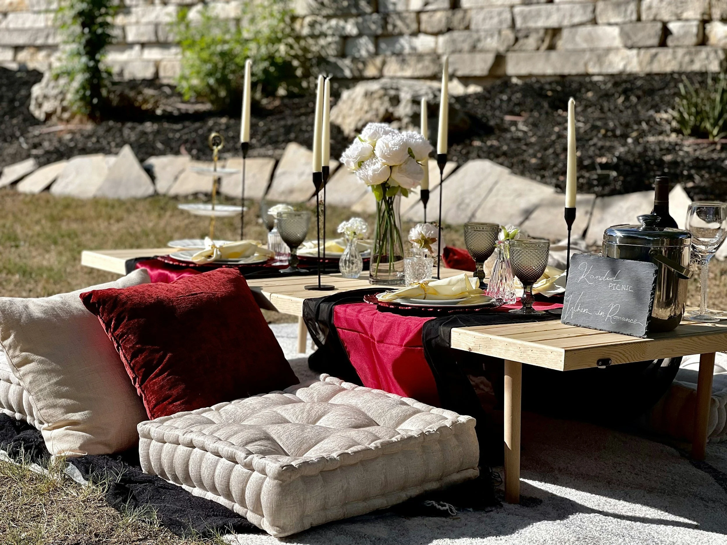 Outdoor picnic table decorated with candles, flowers, and tableware, set on a grassy area with pillows on the ground for sitting.