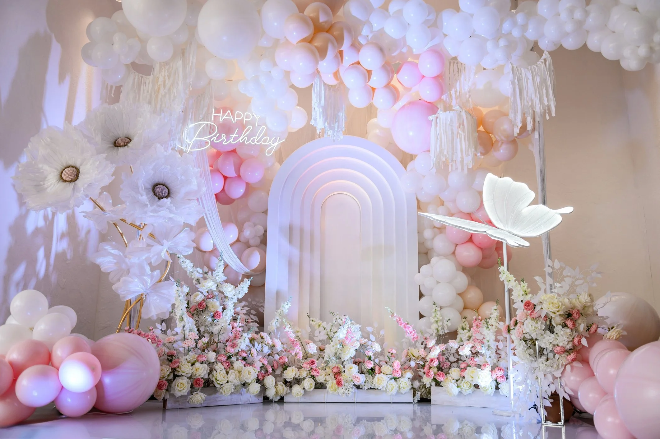 Decorative birthday celebration backdrop with white and pink balloons, white flowers, a rainbow-shaped light display, and a neon sign that says 'HAPPY Birthday'. There is a large white butterfly and floral arrangements at the bottom.