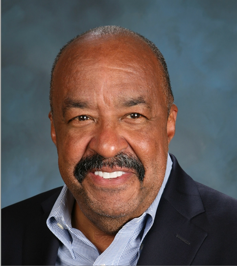 Close-up portrait of an older African American man with a mustache, wearing a navy blazer and light blue checkered shirt, smiling against a blue-gray background.