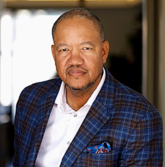 Smiling man in a suit, glasses, and a blue tie, against a gray background.