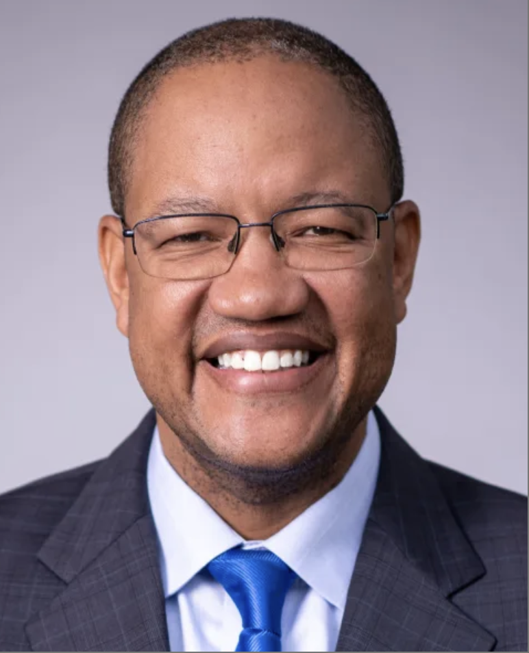 Smiling man in a suit, glasses, and a blue tie, against a gray background.