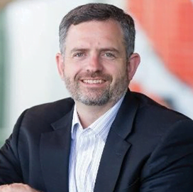 A man with short dark hair and a beard, wearing a dark blazer and a white collared shirt, smiling at the camera in a professional setting.
