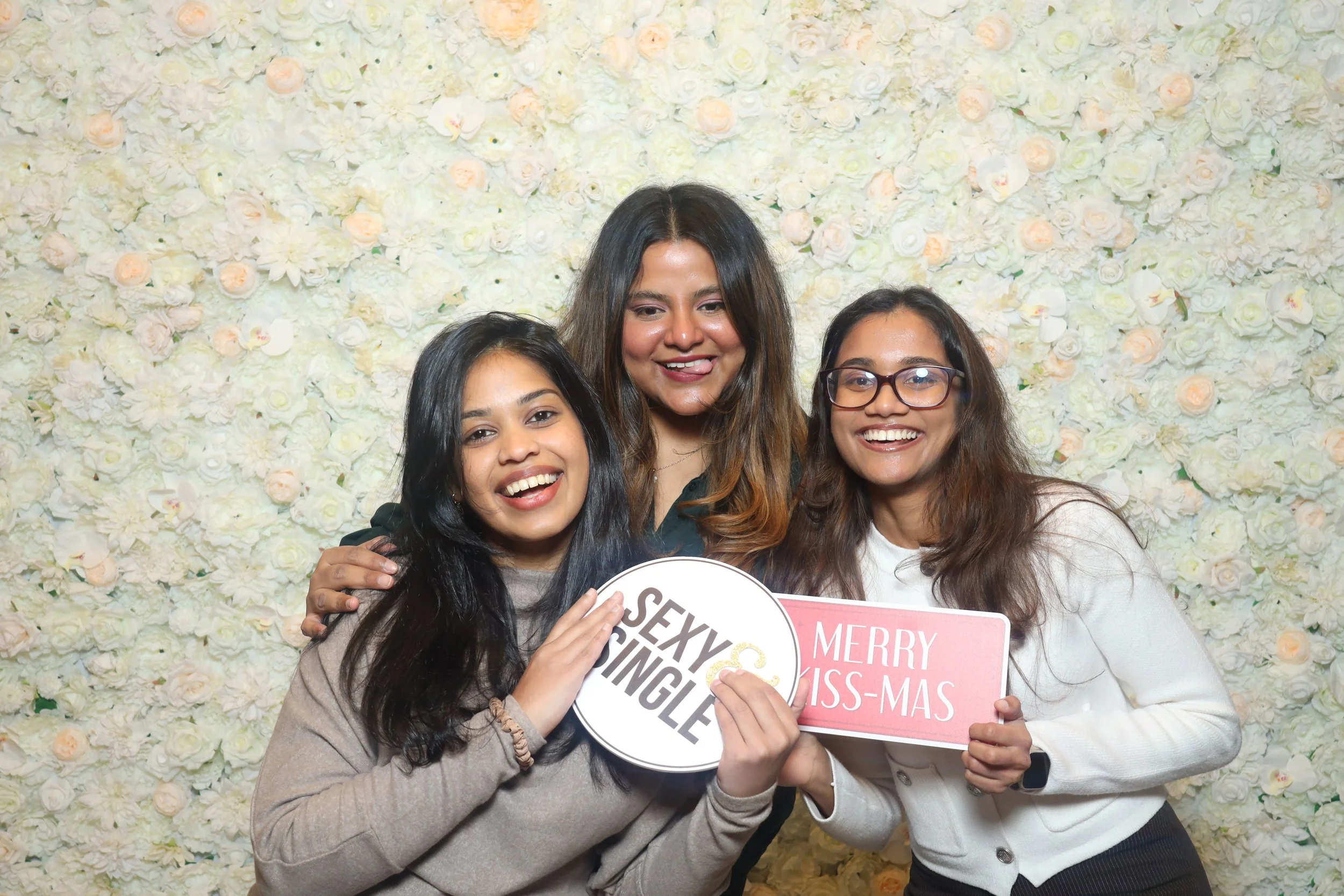 Three women smiling and posing together in front of a white floral backdrop, holding signs that say 'Sexy & Single' and 'Merry Christmas'.