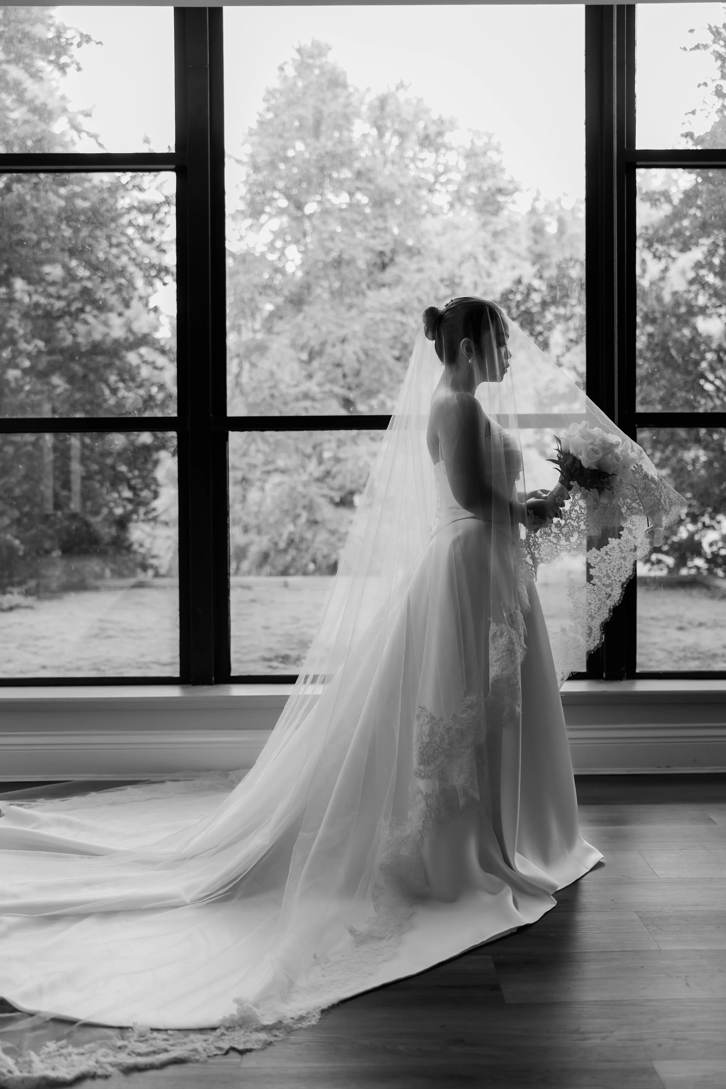 Black and white photo of a bride holding a bouquet, standing by a large window, with a scenic outdoor view of trees.