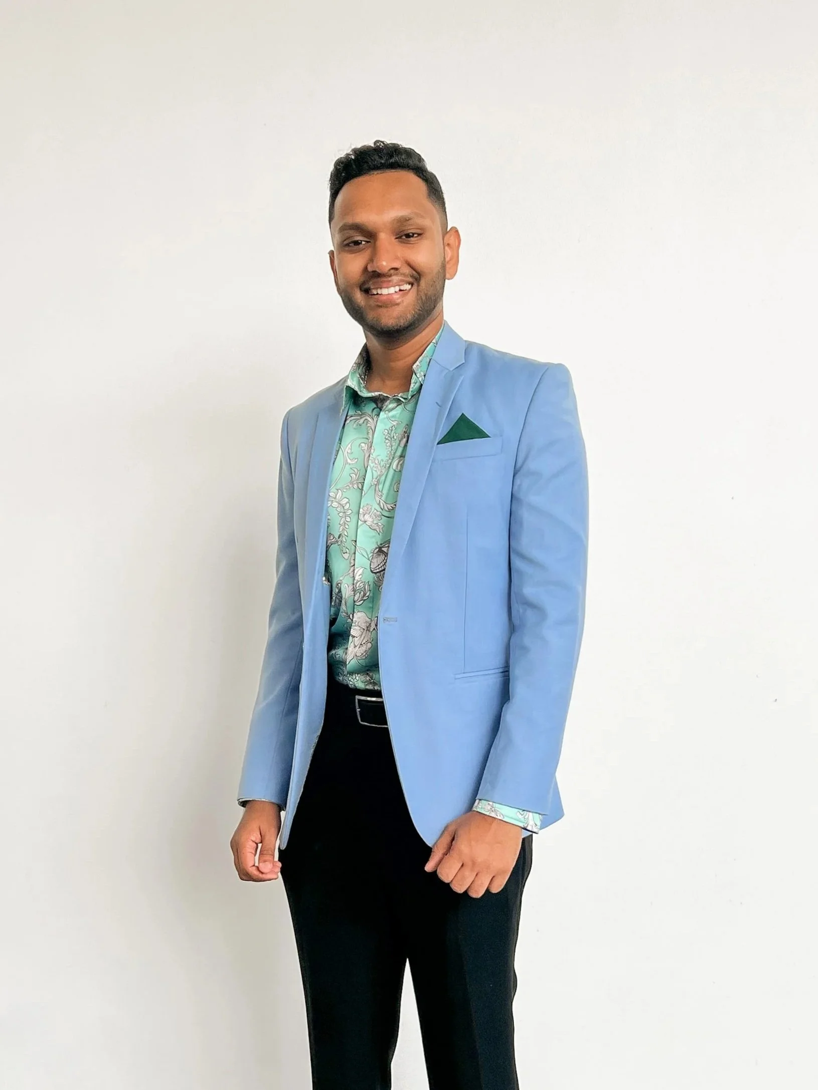 A man in a light blue blazer, patterned shirt, and black trousers standing against a plain white background, smiling at the camera.