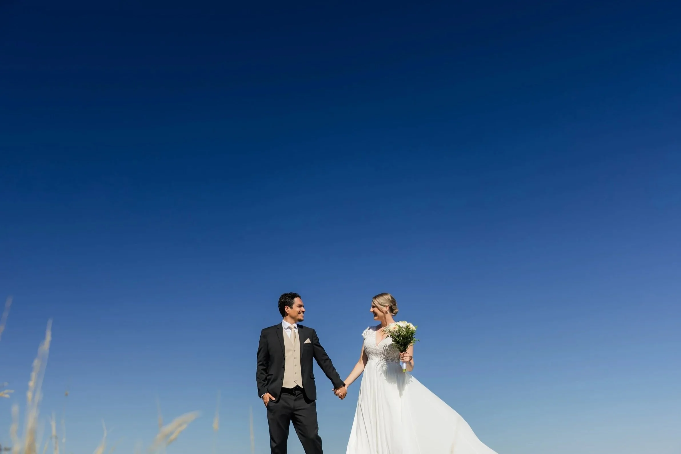 A bride and groom standing outdoors, holding hands and smiling at each other, against a bright blue sky. The bride is holding a bouquet of flowers and wearing a white wedding dress, while the groom is dressed in a black suit with a beige vest and tie.
