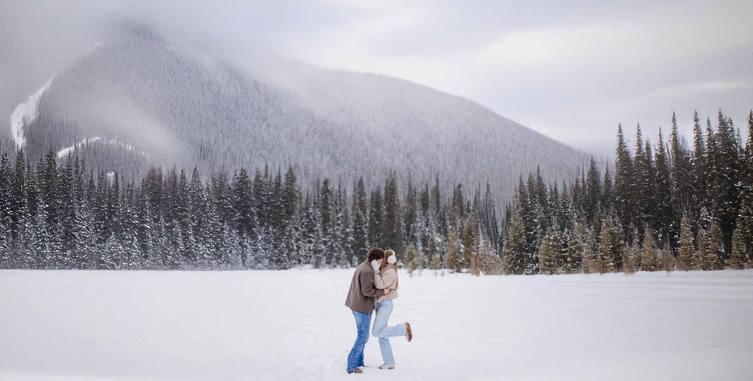 A couple shares a kiss in a snowy landscape with mountains and a forest in Vancouver
