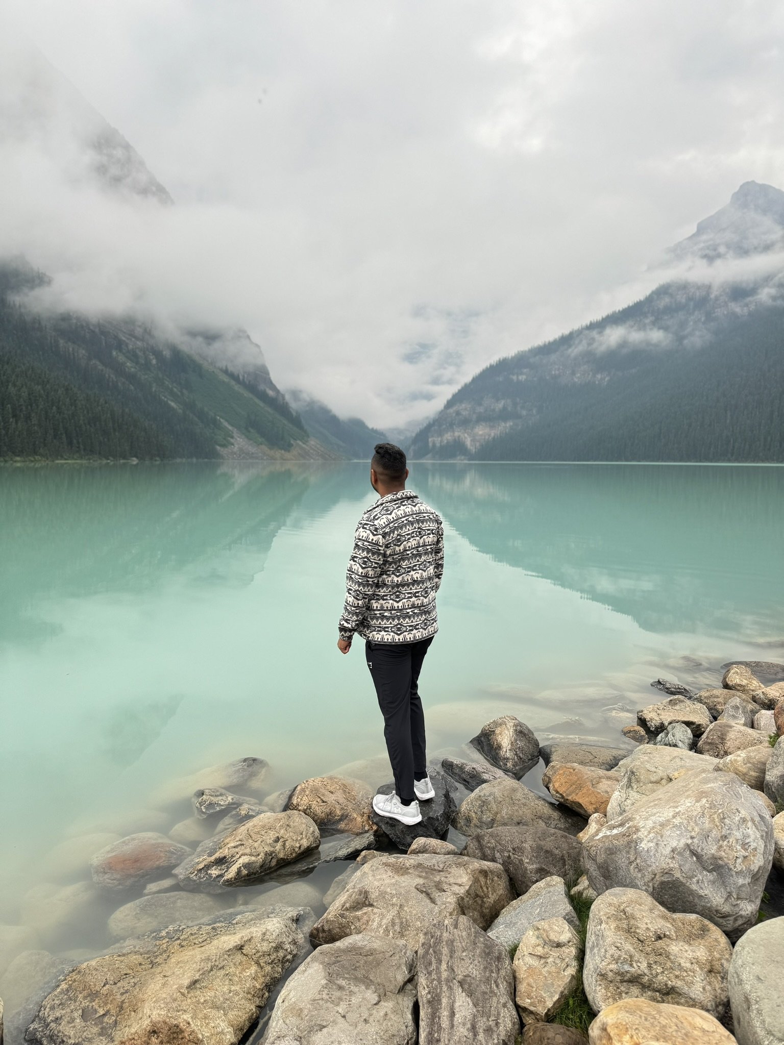 A person standing on rocks by a lake, looking out at mountains and cloudy sky.
