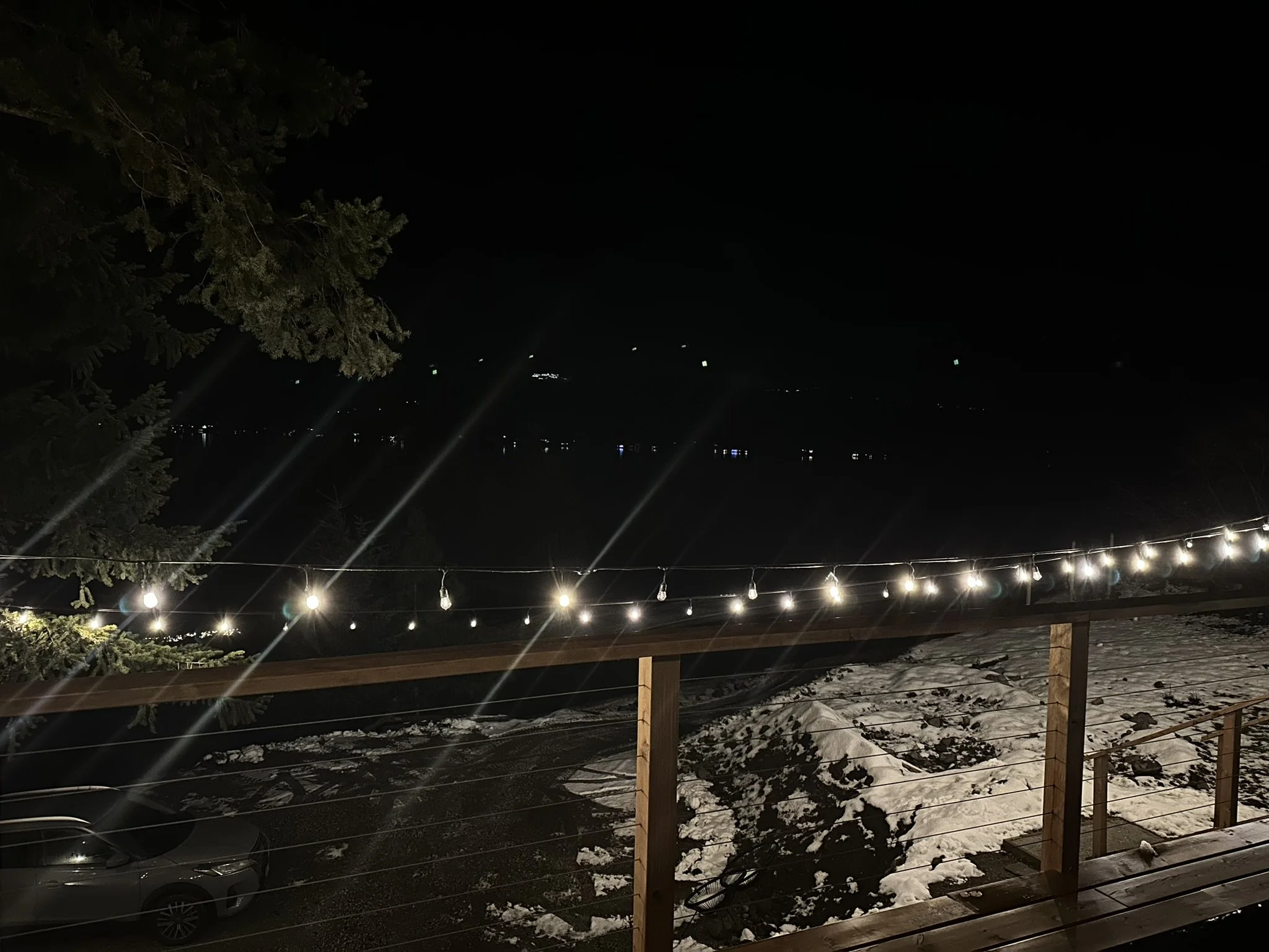 Nighttime outdoor view with string lights on a wooden deck, snow on the ground, and a dark lake with faint lights reflecting on the water in the distance.