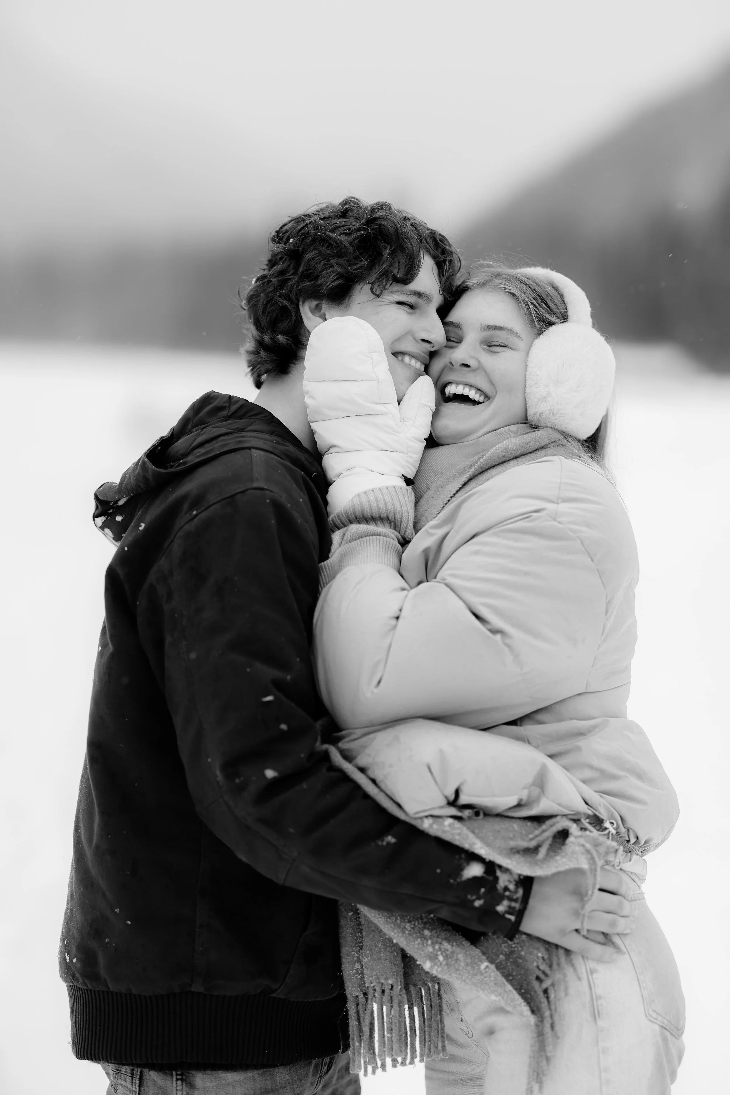 A black and white photo of a young couple embracing outdoors in winter, smiling and laughing, with snow on their clothing and a blurred snowy landscape in the background.