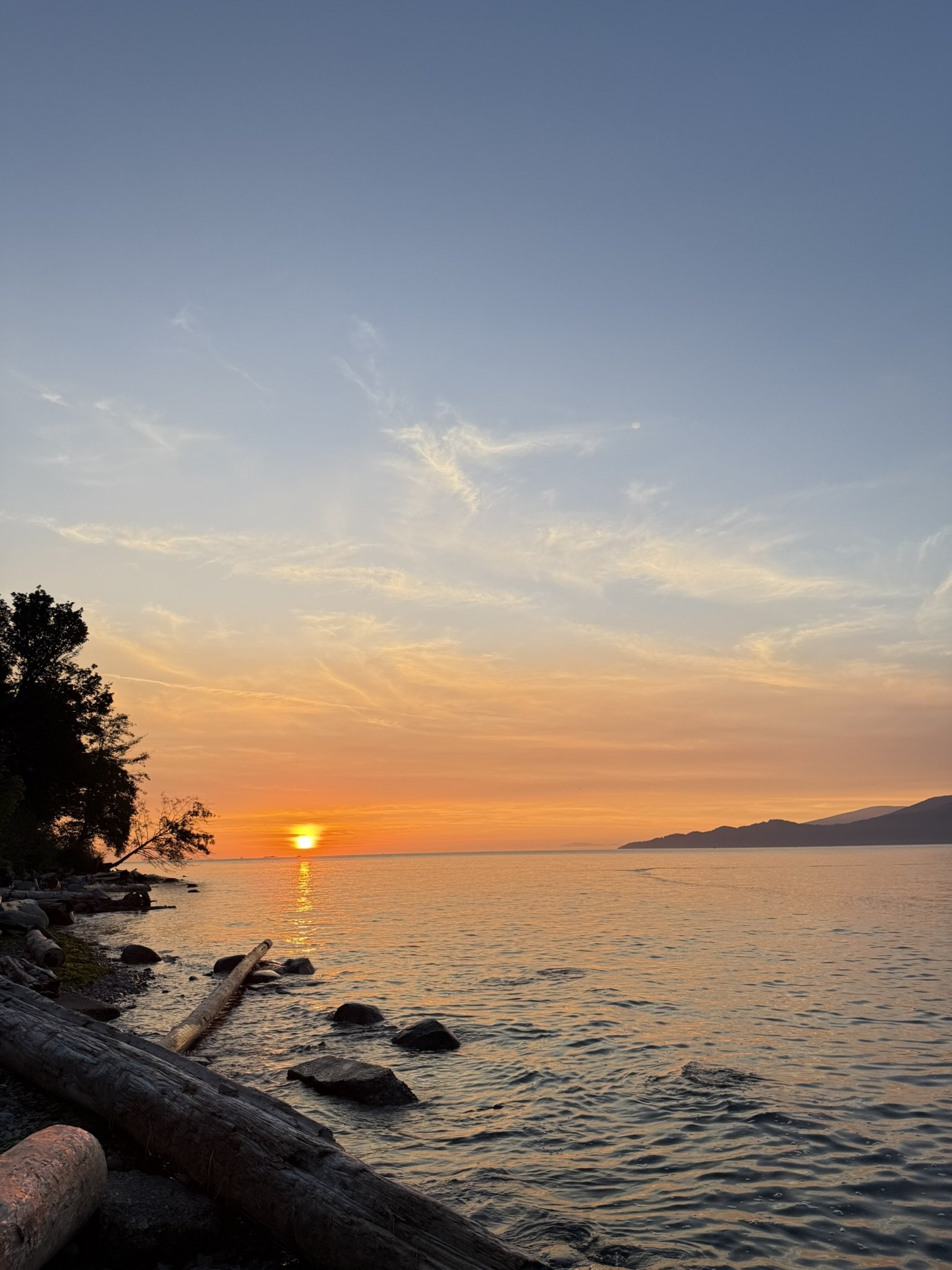 Sunset over a calm body of water with a colorful sky and distant mountains in the background, with rocks and logs along the shoreline.