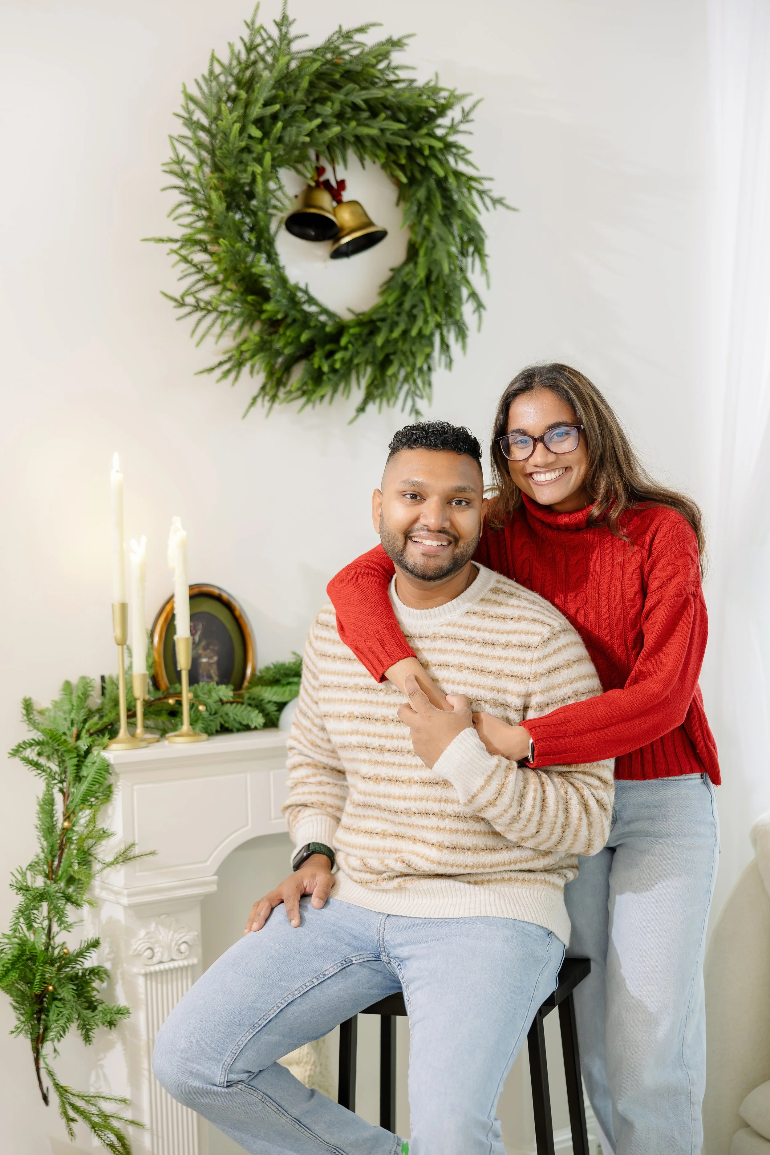 A smiling couple celebrating Christmas in their home, decorated with a green wreath with bells hanging on the wall, a white fireplace with candles and greenery, and a small Christmas tree branch, wearing cozy sweaters.