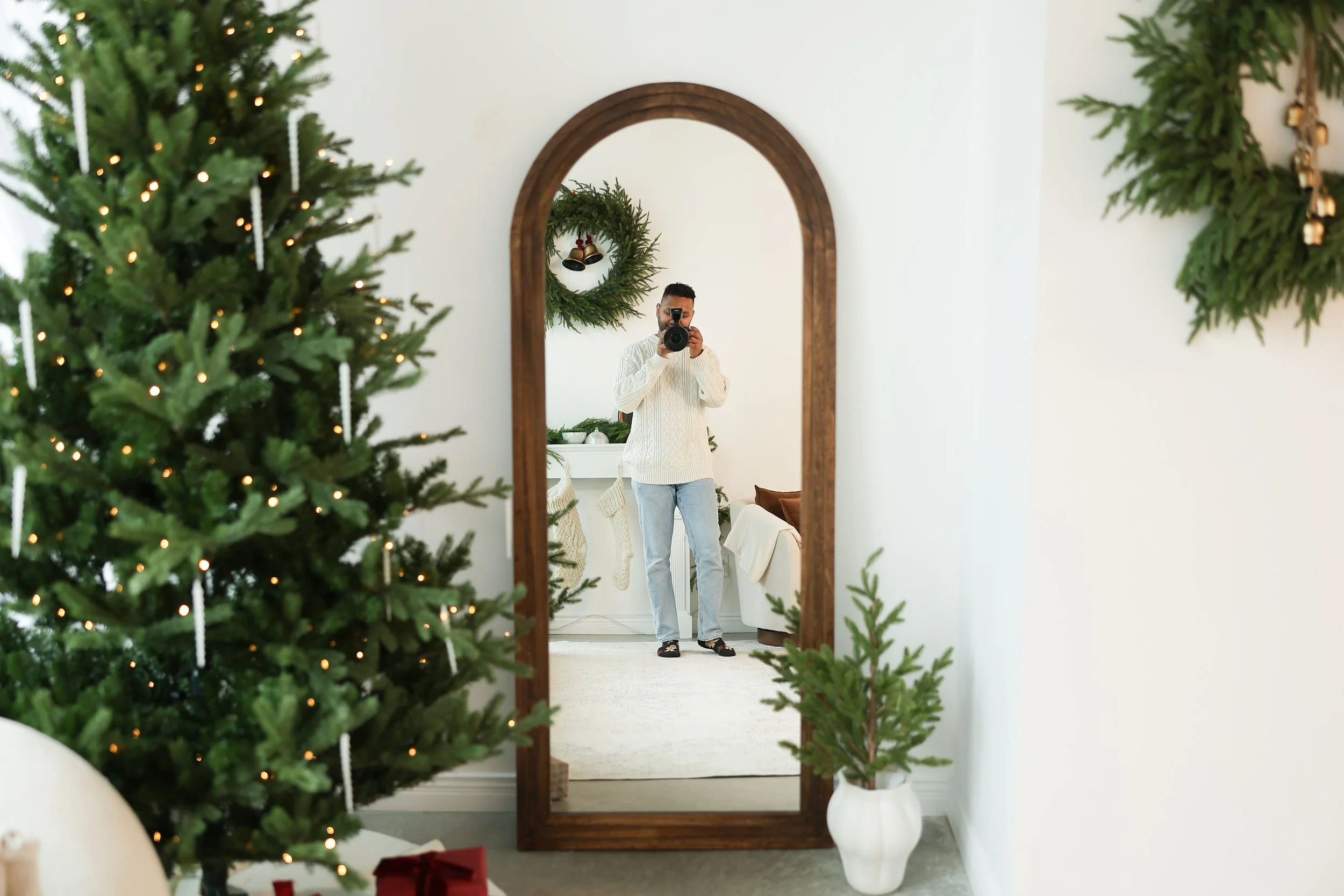 A person taking a photo of their reflection in a large freestanding mirror decorated for Christmas, with a Christmas tree and holiday decorations visible in the room.