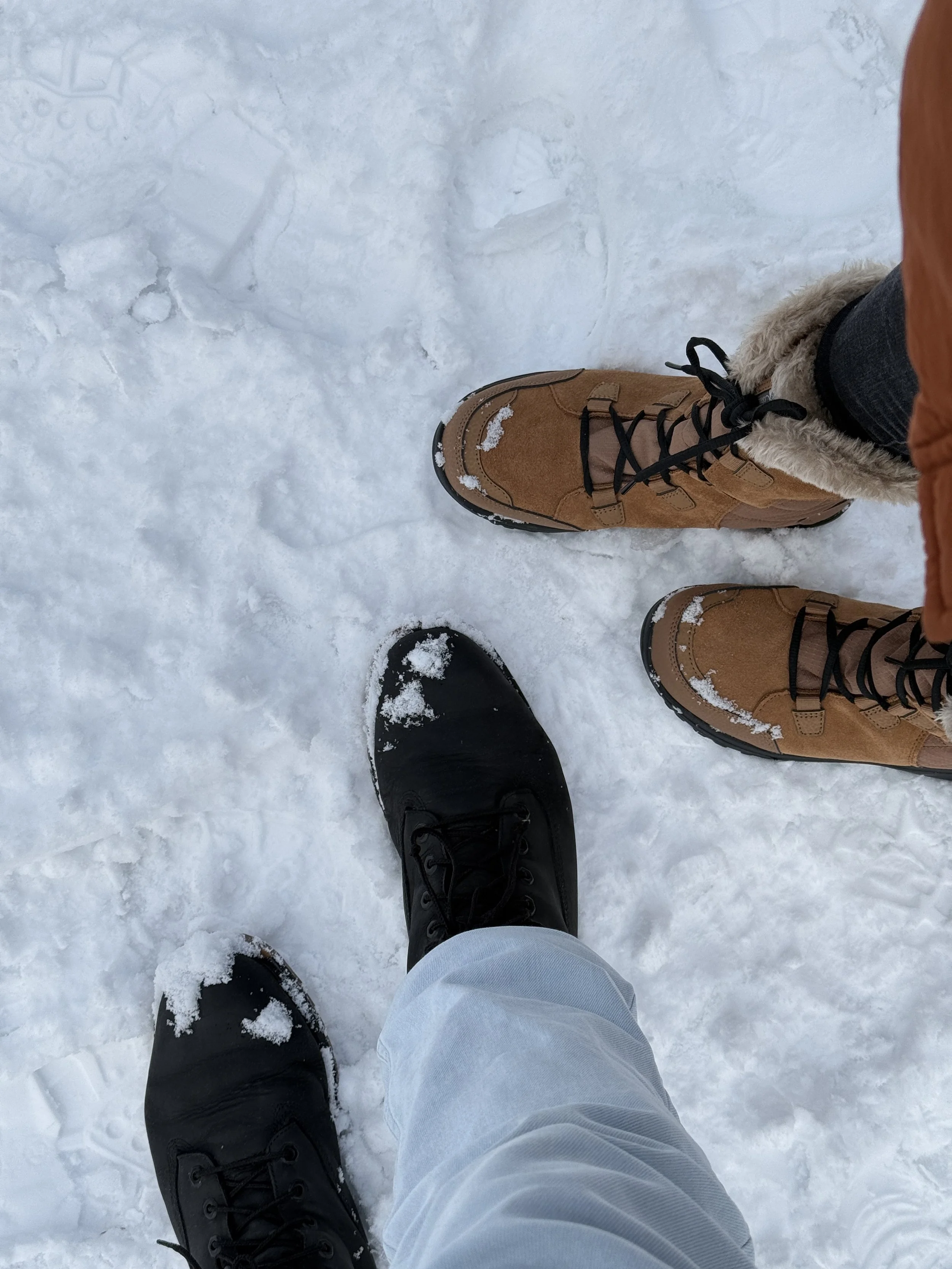 Two pairs of winter boots, one black and one tan, stand on snow. A person in black shoes and light-colored pants is in the foreground, while another person in tan boots and dark pants is beside them.