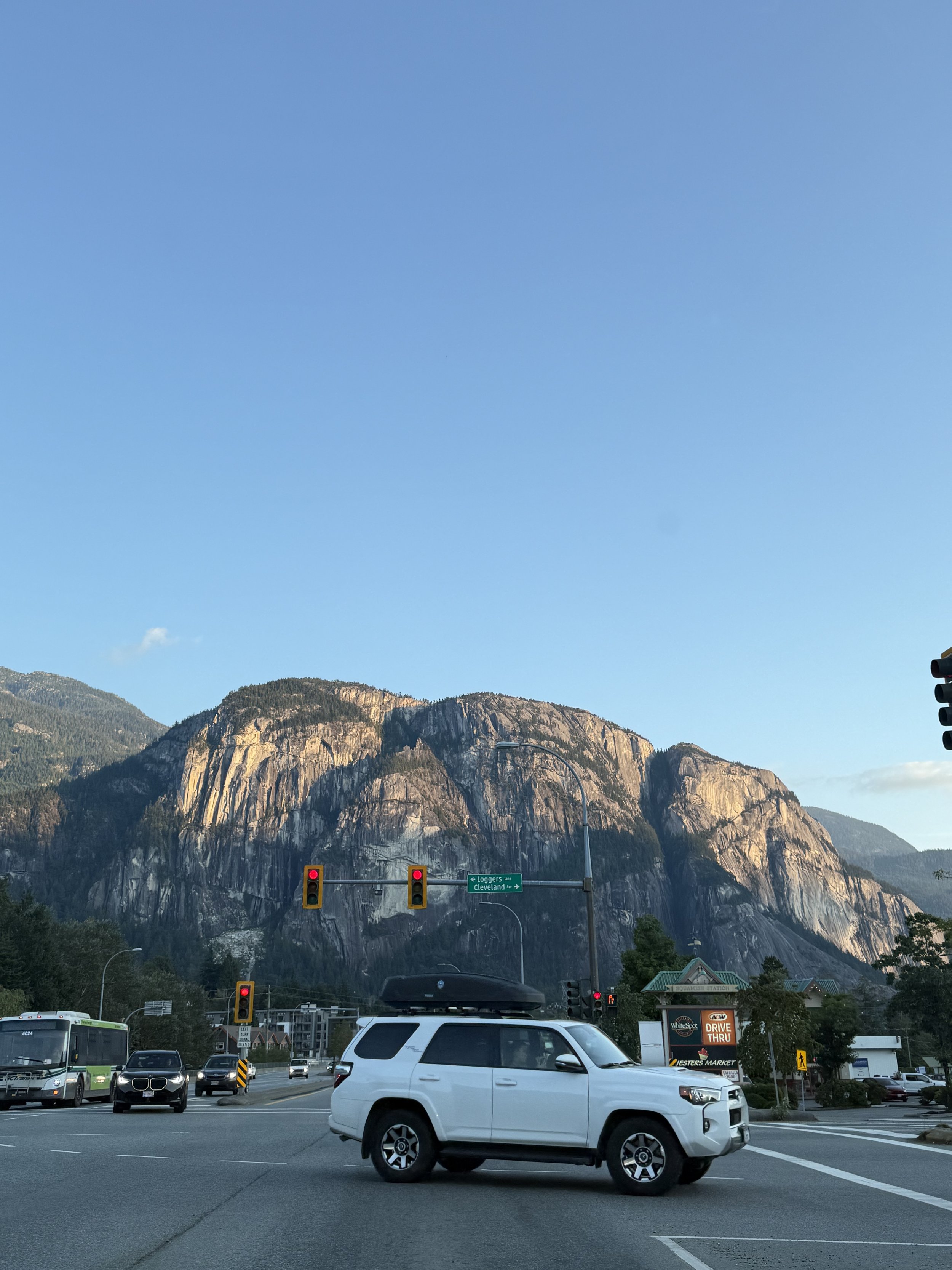 A busy intersection with several vehicles, including a white SUV, a bus, and a black car, with a mountain in the background and traffic lights showing red