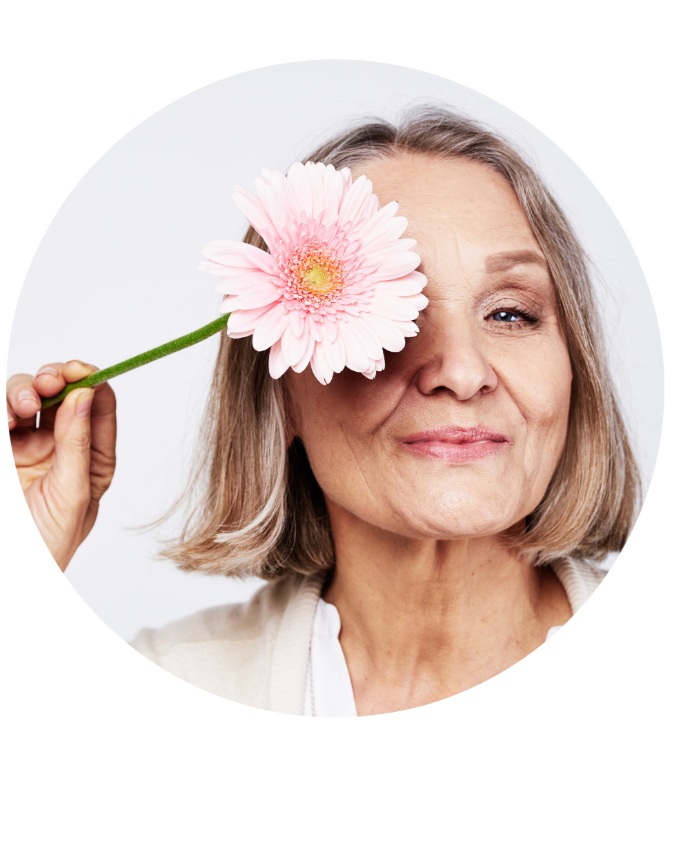 An elderly woman with shoulder-length gray hair holds a pink gerbera flower over her eye, smiling softly against a plain white background. Aging Gracefully. Anti-aging