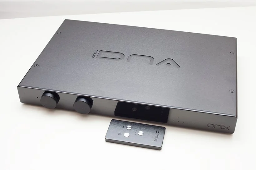 A black Onix audio amplifier with two knobs and a small remote control placed in front of it. ONIX DNA Range Amplifiers -  (C)  www.oa21.com