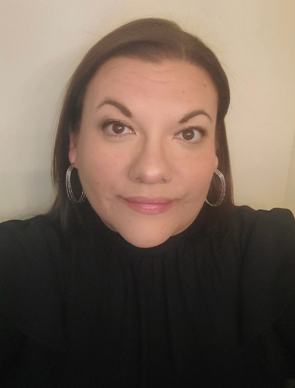 A woman with brown hair and hoop earrings smiling for a selfie against a plain wall.