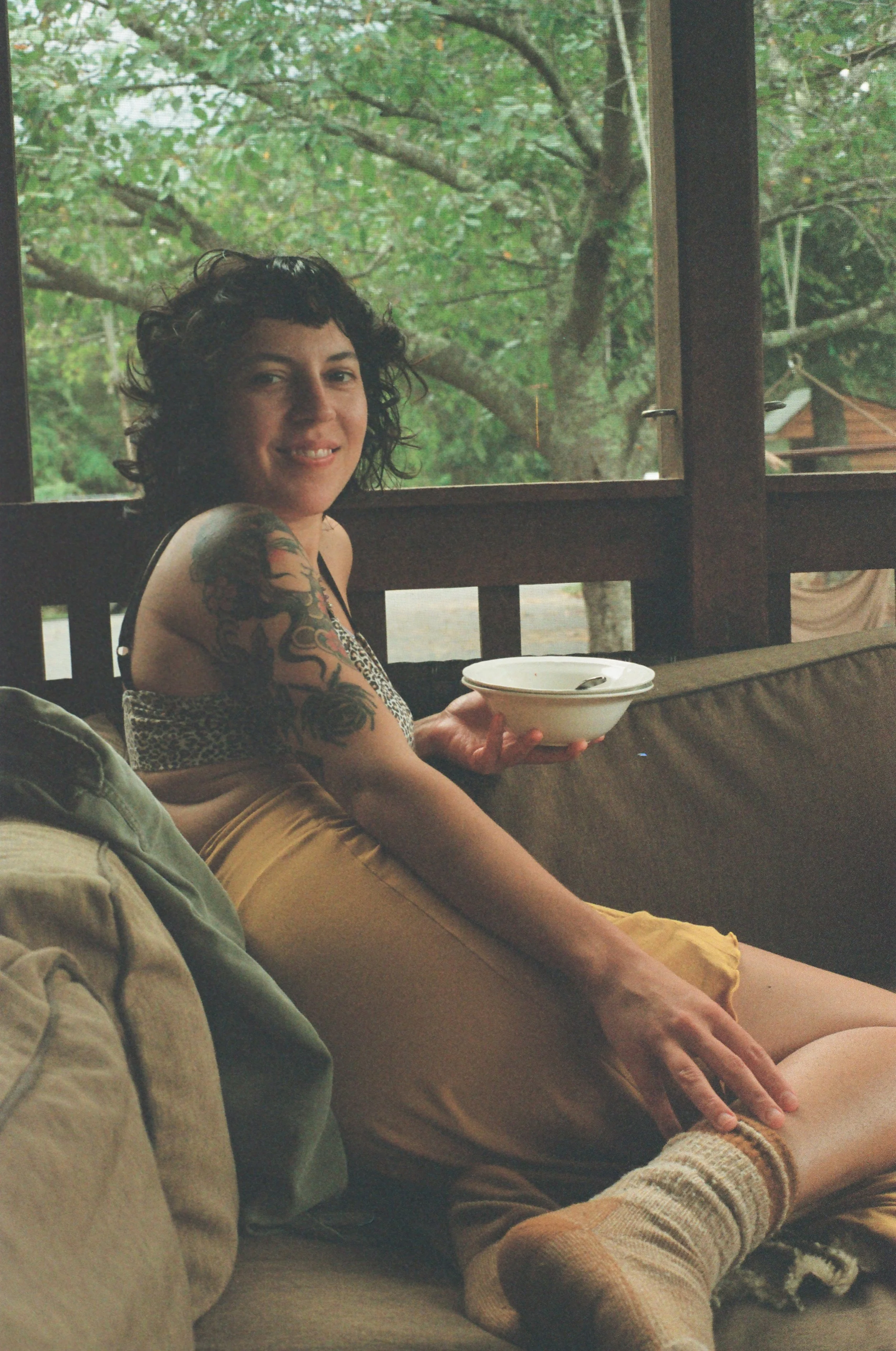 A woman with dark curly hair and tattoos on her arm, sitting on a sofa with a bowl and spoon, smiling at the camera, with a window showing trees outside in the background.