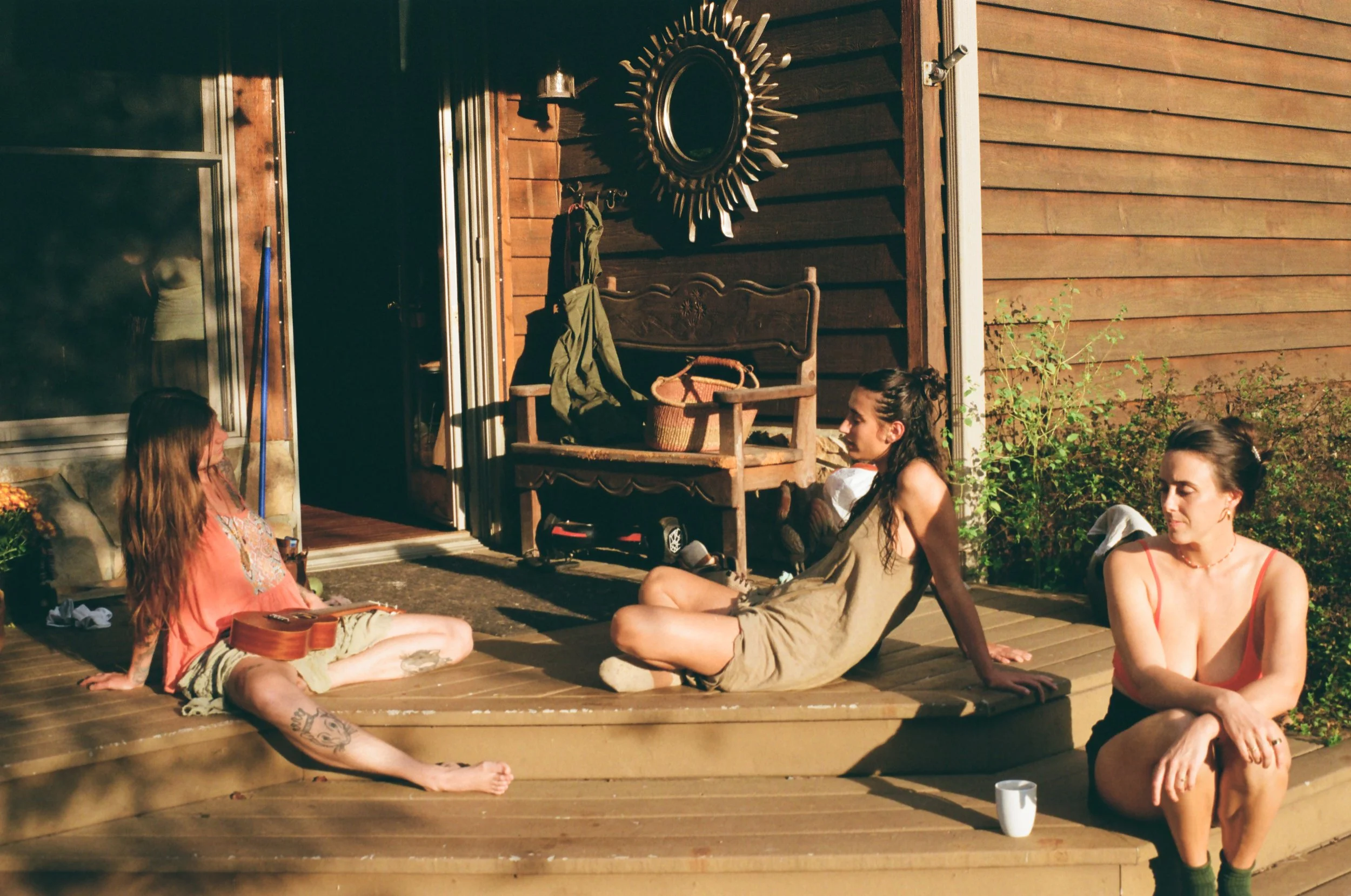 Four women sitting on a wooden porch in front of a house, engaged in conversation, with warm sunlight illuminating the scene.