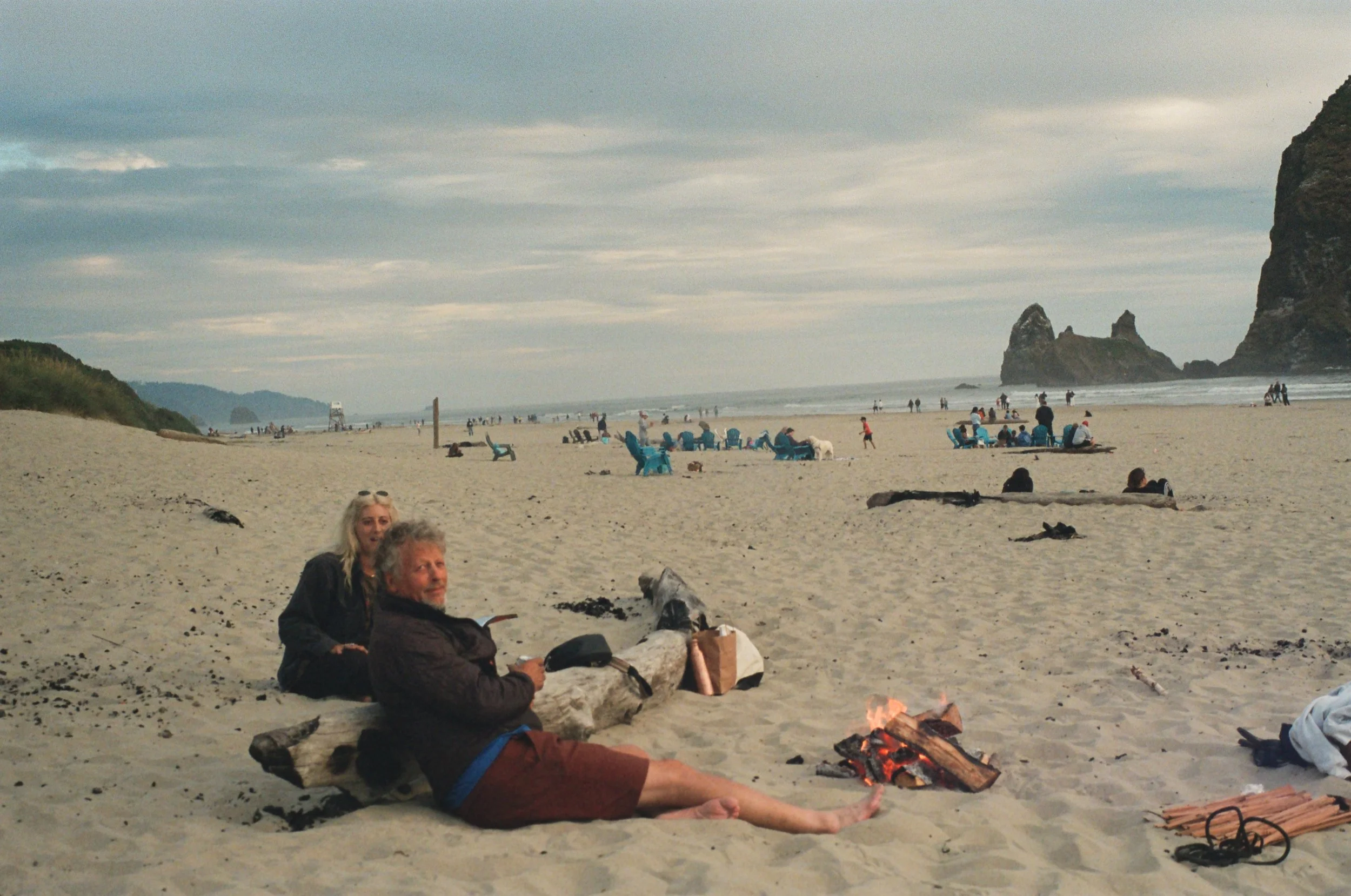 Beach scene with people sitting around a campfire, some lying on the sand, and others sitting on beach chairs, with large rock formations and the ocean in the background.