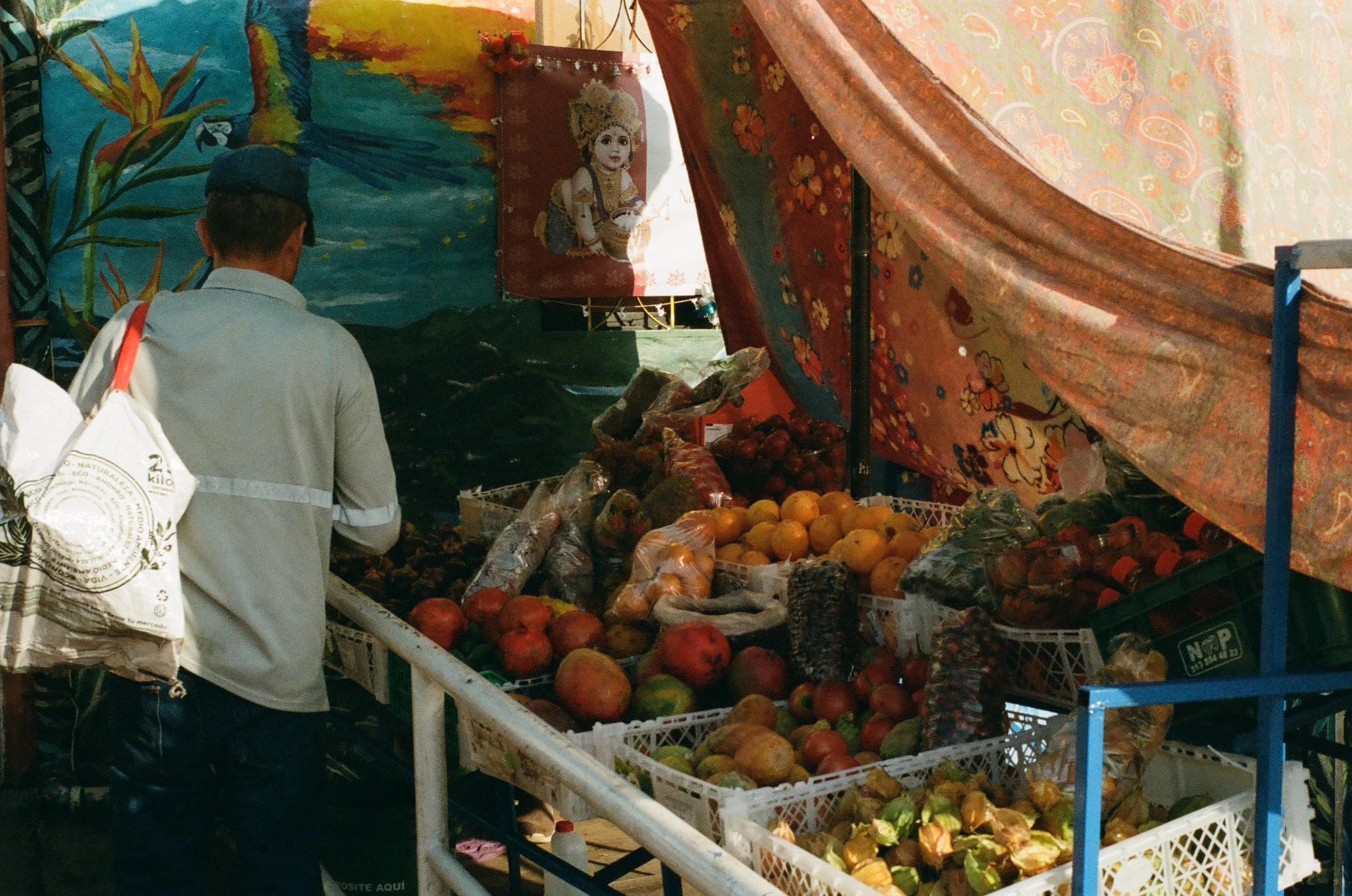A person shopping at a fruit stand with a canopy decorated with colorful paintings and images of children, displaying various fruits like pomegranates, oranges, and avocados.