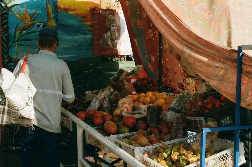 Market stall selling various fruits including pomegranates, apples, and oranges, with colorful fabric curtains overhead and a painted mural in the background.