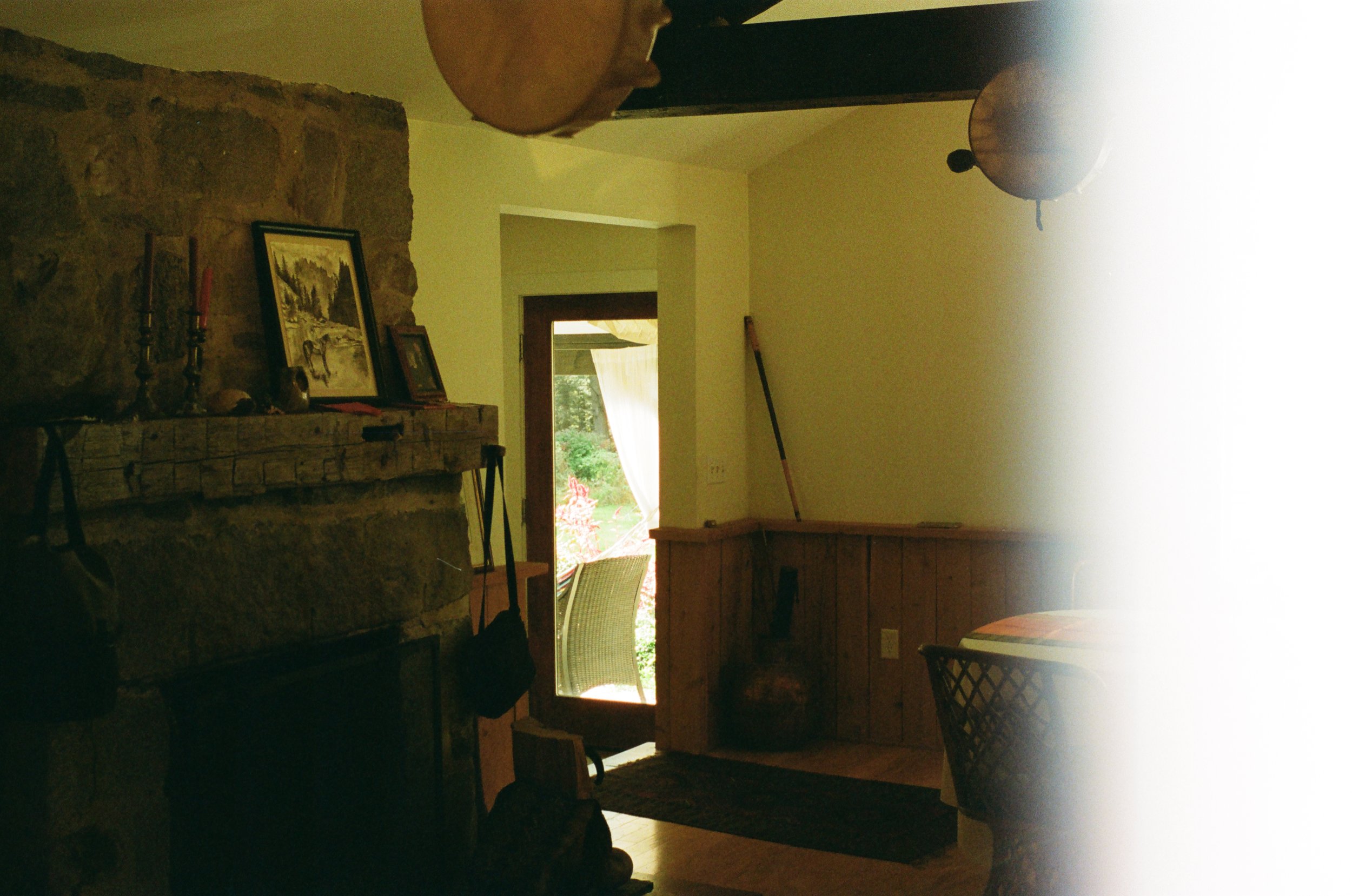 Interior of a rustic living room featuring a stone fireplace with framed pictures and candles, a bistro table with chairs, wooden wall paneling, and an open door leading to a garden.