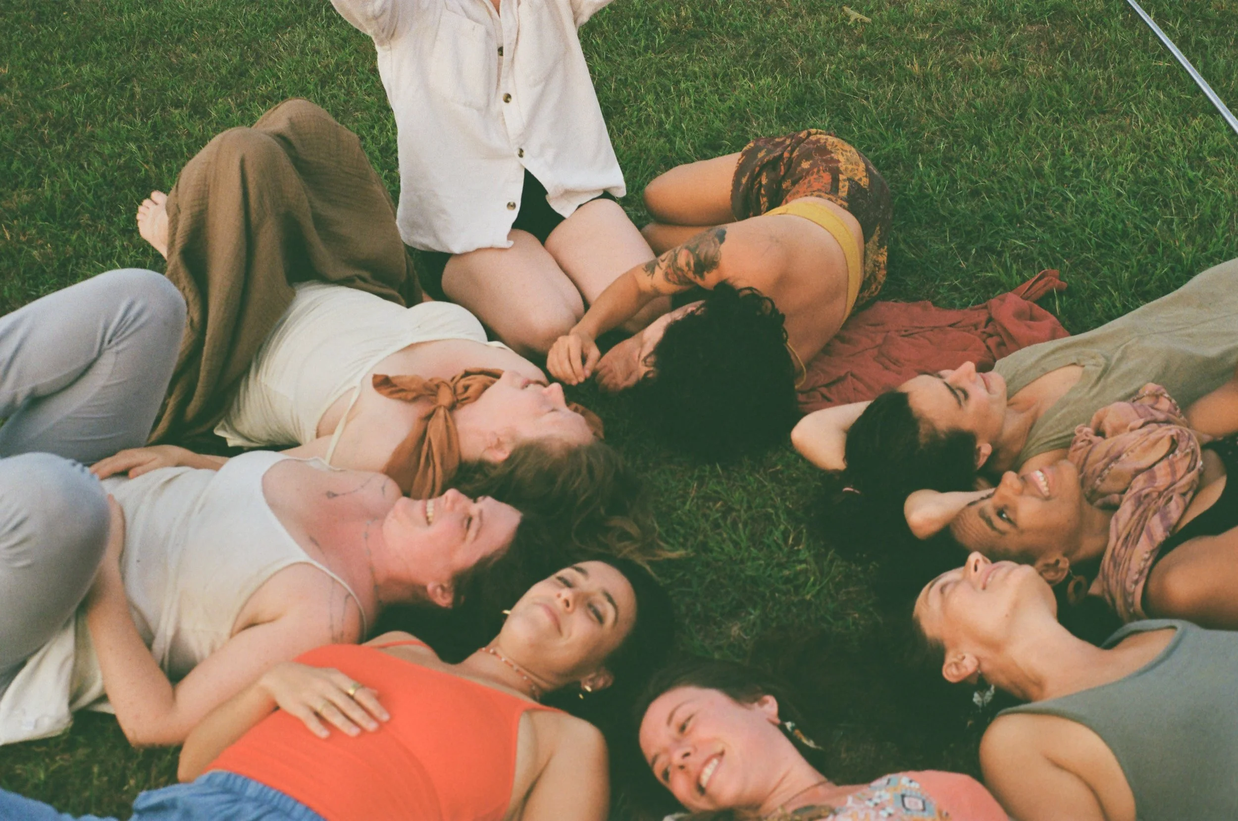Several women lying on grass, smiling, and looking relaxed. Some are lying with heads near each other, forming a circle, while others are positioned on the grass, with one woman sitting up.