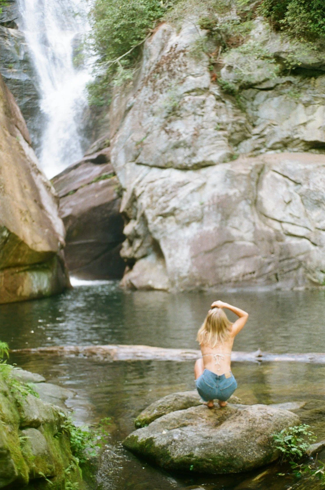 A woman with long blonde hair crouches on a large rock in a river, facing a waterfall cascading down rocks surrounded by green foliage.