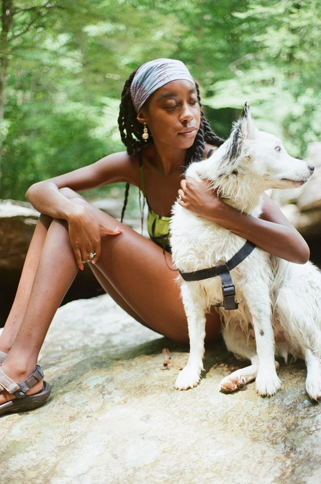A woman sitting on a rock in a green forest, holding a white dog with blue eyes, both looking to the right.