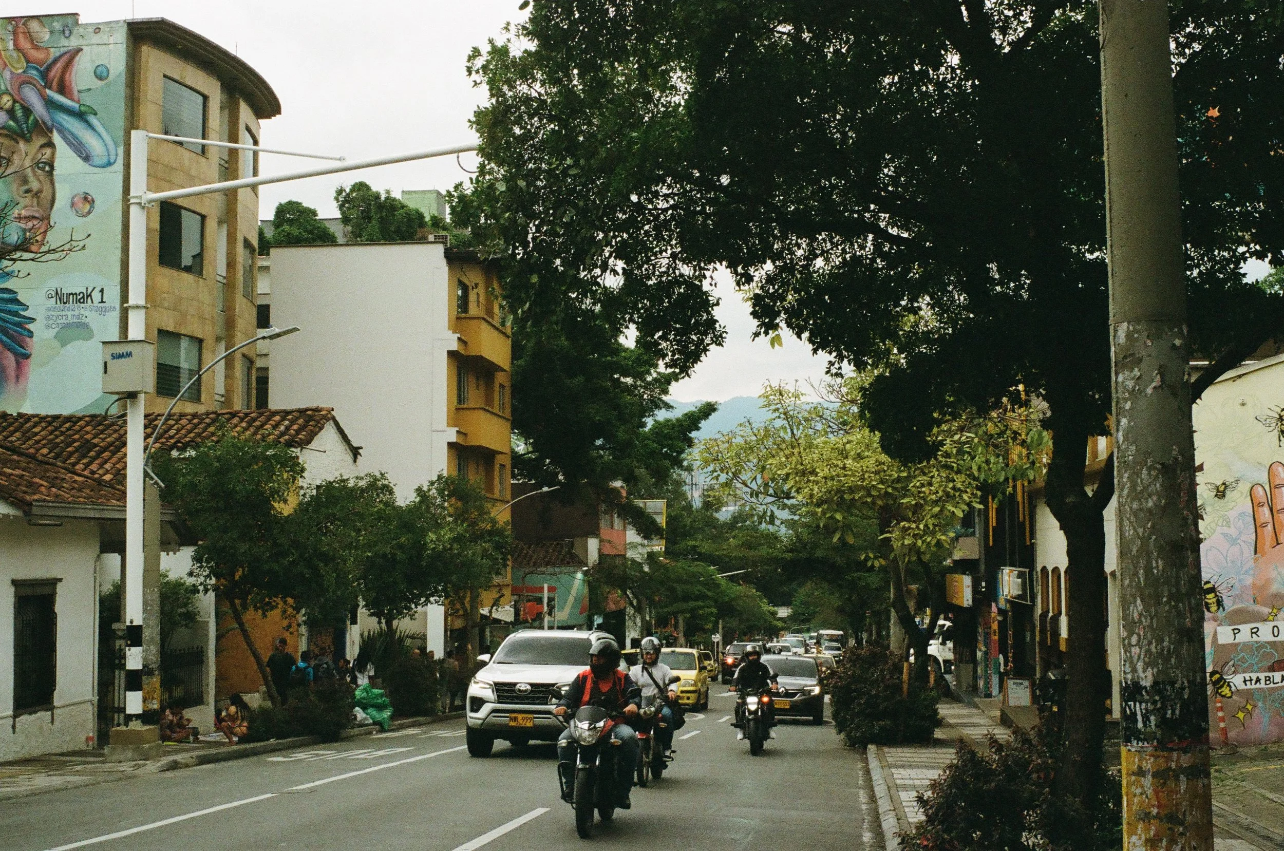 A city street with cars and motorcycles, shops, trees, and colorful building murals.