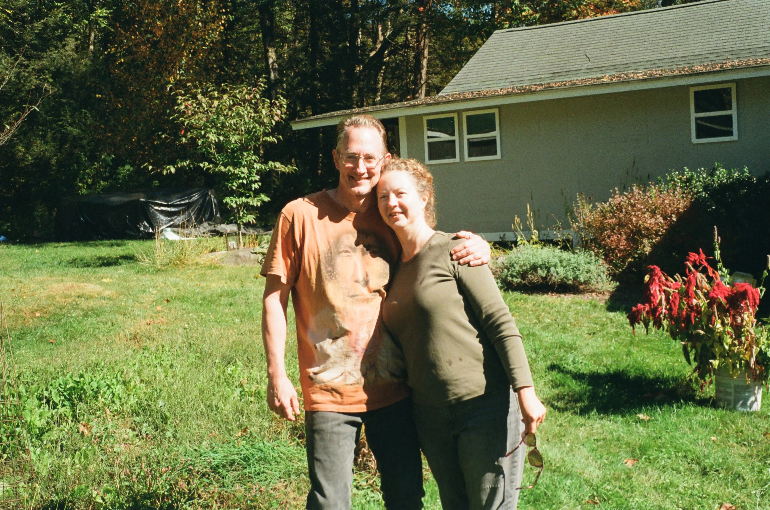 A man and woman standing close together outdoors in a yard, smiling and embracing each other. The man is wearing a graphic t-shirt and the woman is in a long-sleeve shirt and holding sunglasses. There are bushes and a house in the background, with tr