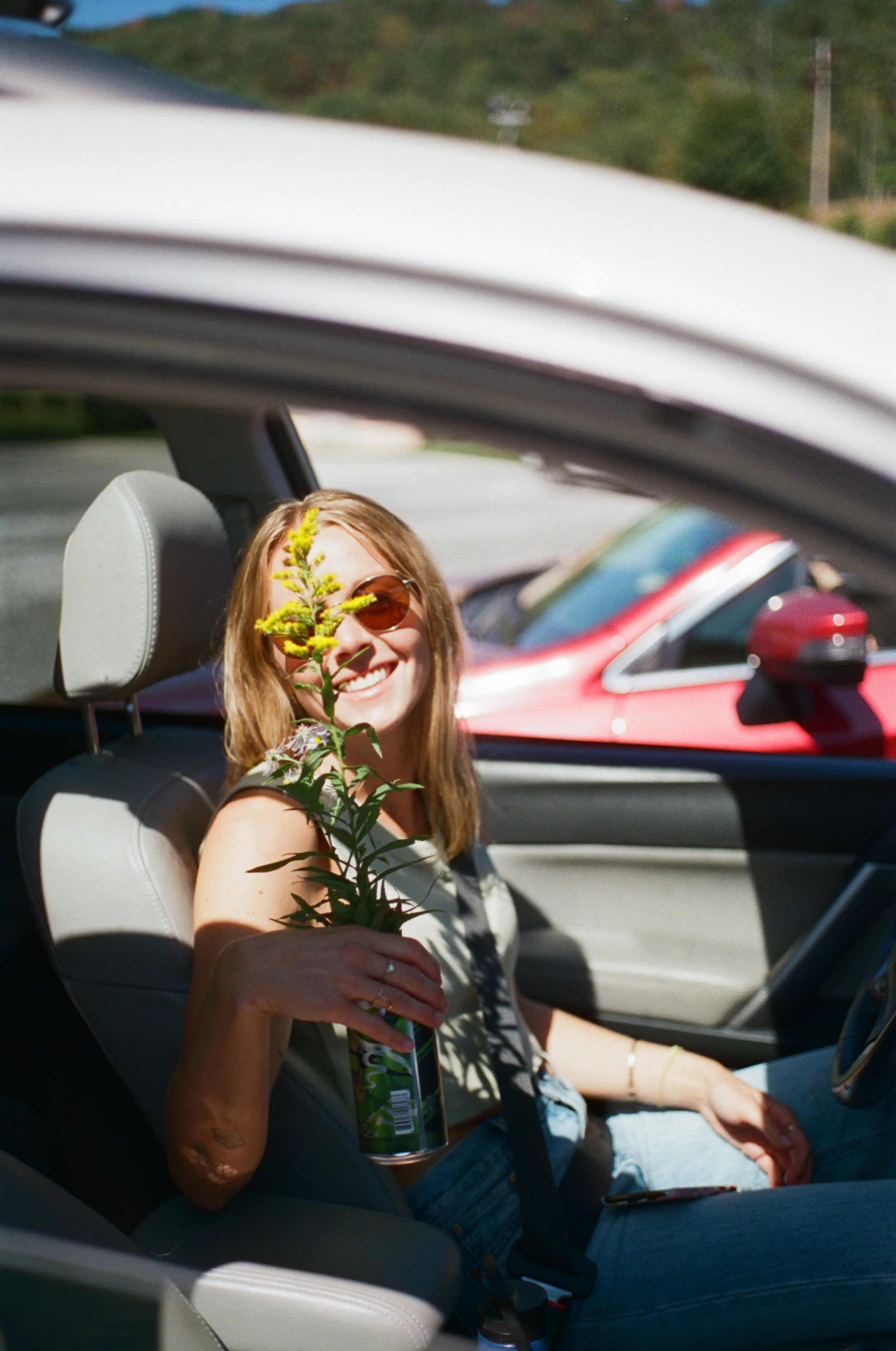 A young woman with brown hair and sunglasses sitting in the passenger seat of a convertible car, holding a green plant with yellow flowers and smiling at the camera.