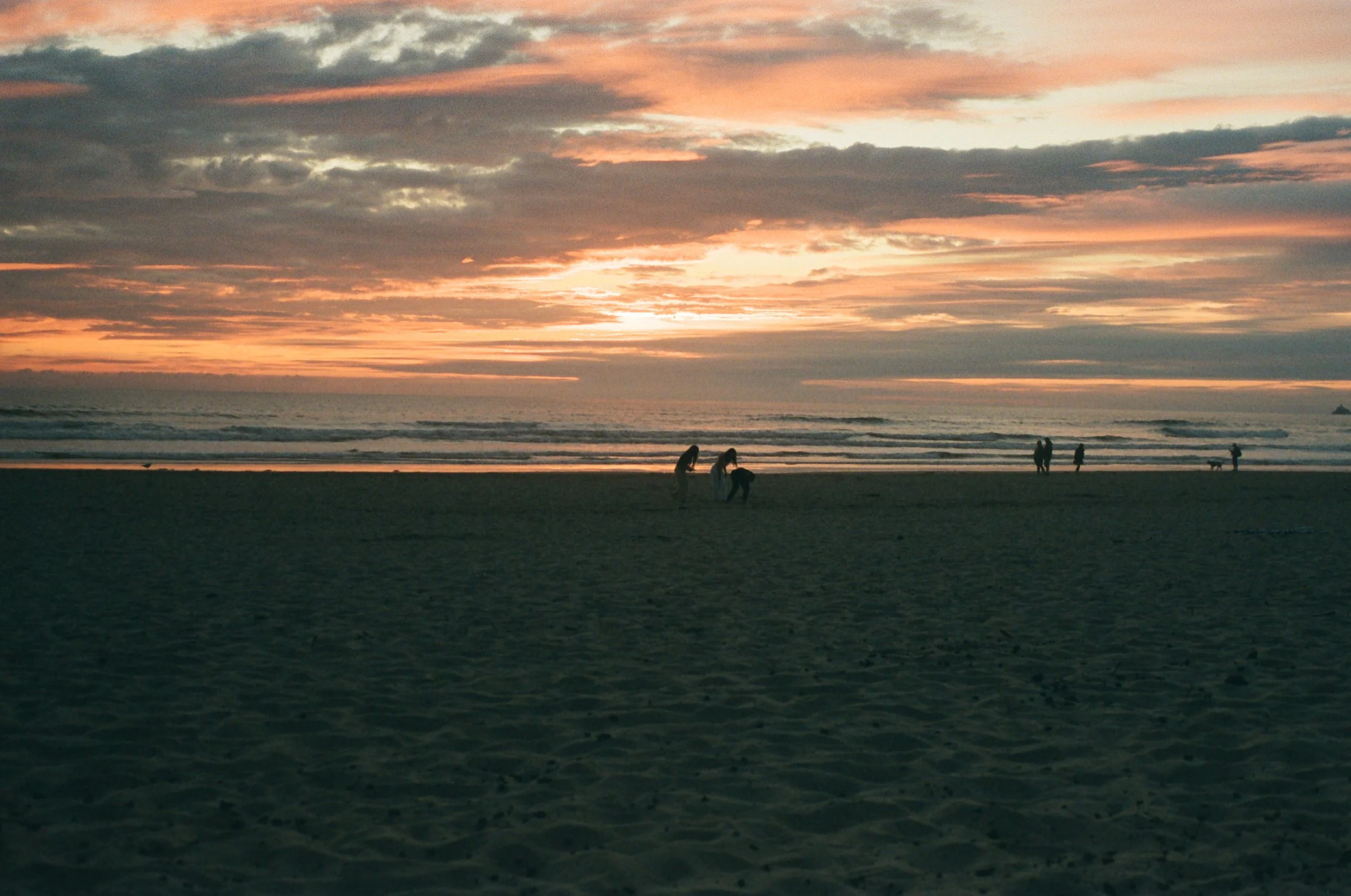 People walking and gathering on a beach at sunset, with clouds in the sky and the ocean in the background.