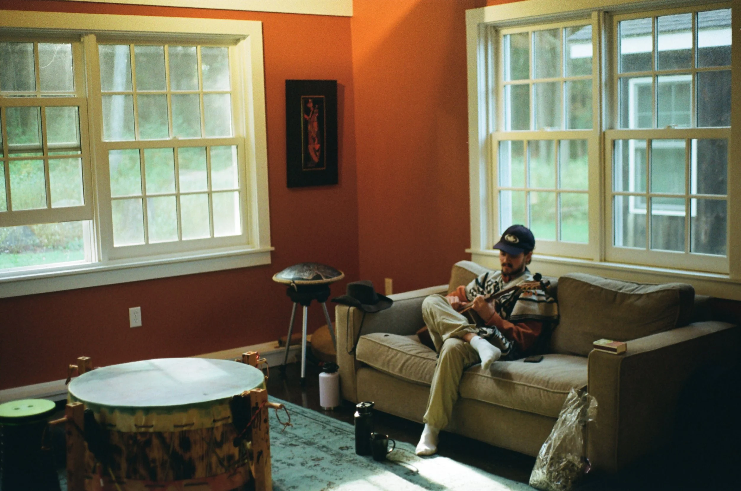 A man sitting on a beige couch playing guitar in a living room with large windows, orange walls, and various objects on the floor and furniture.