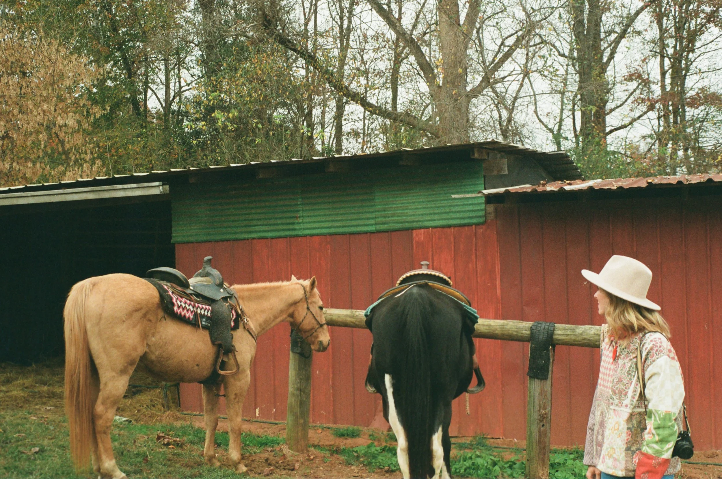A woman in a white wide-brimmed hat wearing a colorful patchwork jacket stands next to a horseshoe in a paddock. Two horses, one with a saddle and the other covered with a blanket, lean over a wooden fence in front of a red barn with a green corrugated metal roof. Trees in the background have autumn foliage.