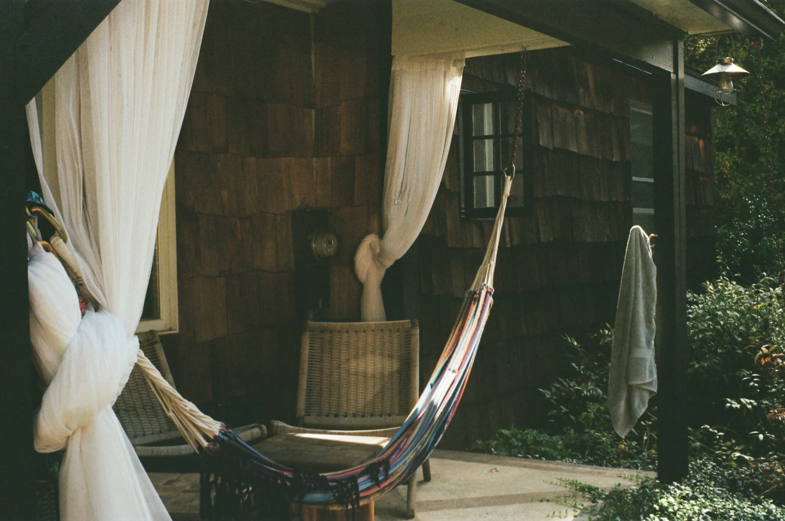 A cozy outdoor porch featuring a hammock, a wicker chair, a hanging towel, and surrounded by greenery.