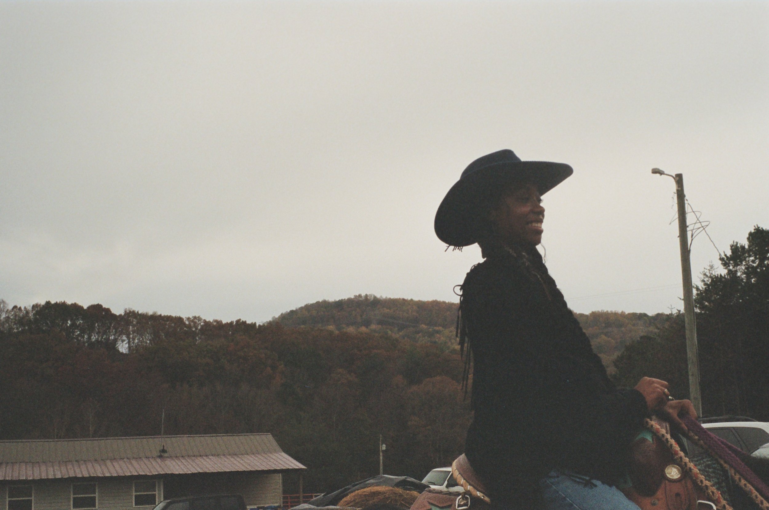 A woman smiling while riding a horse outdoors, wearing a large black hat and a black jacket, with trees and a hill in the background.