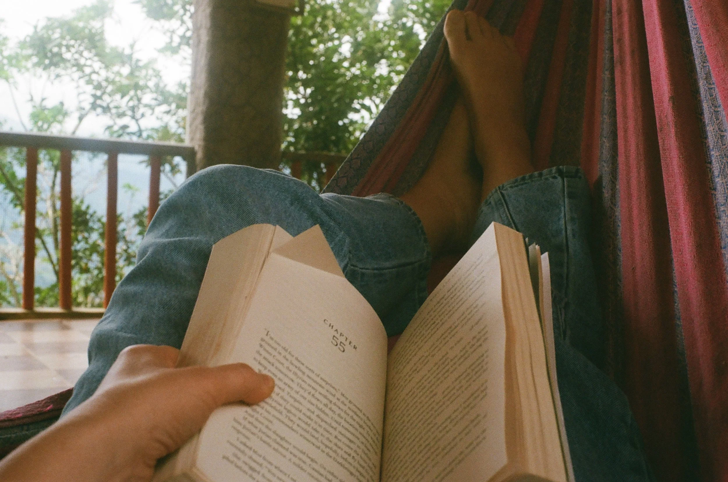 Person sitting outdoors, reading a book, with legs stretched on a hammock, trees in the background.