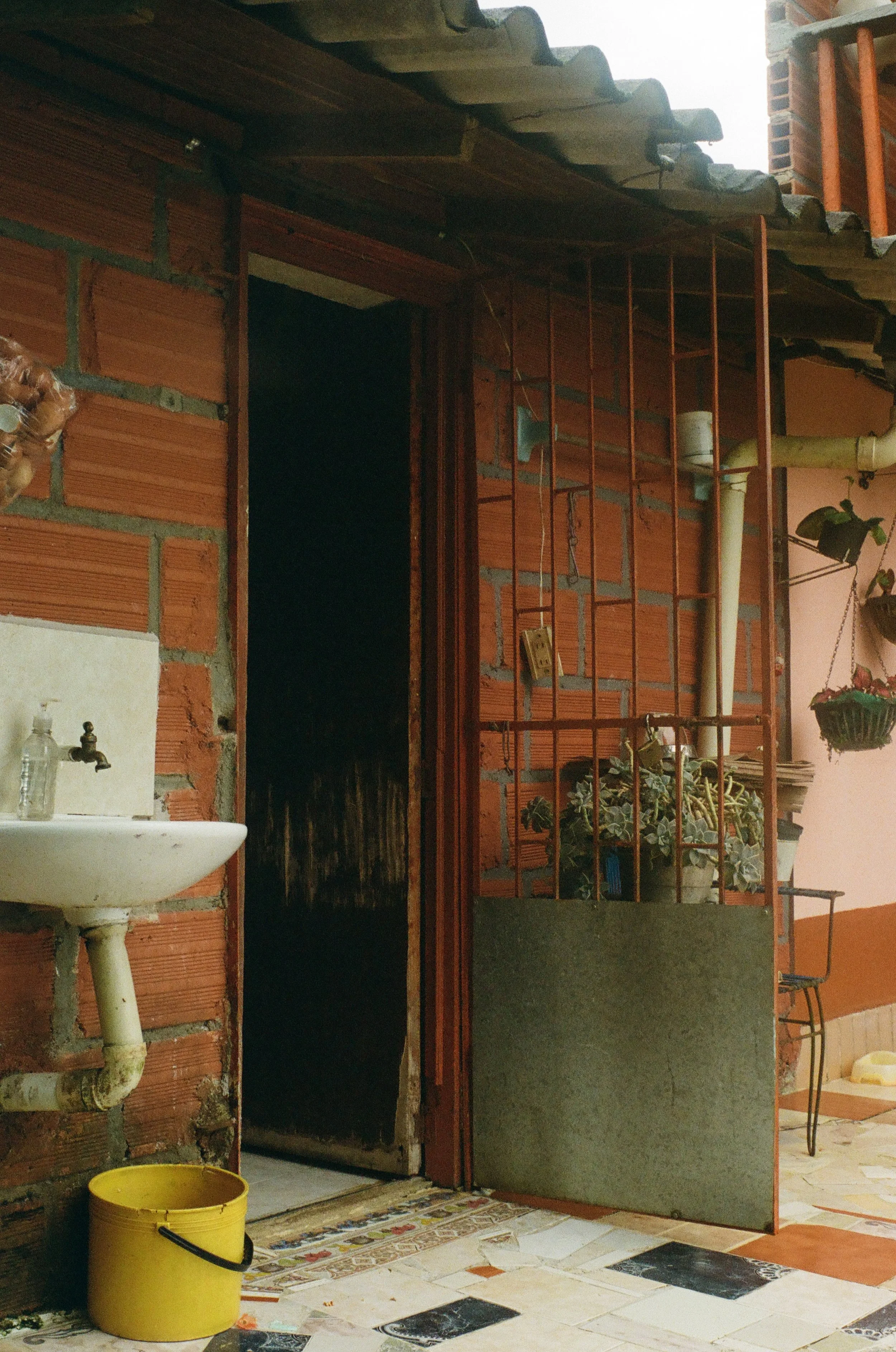 Exterior view of a brick wall with a doorway, small sink with a soap dispenser, potted plants on a metal stand, hanging plants, and a yellow bucket on tiled floor.