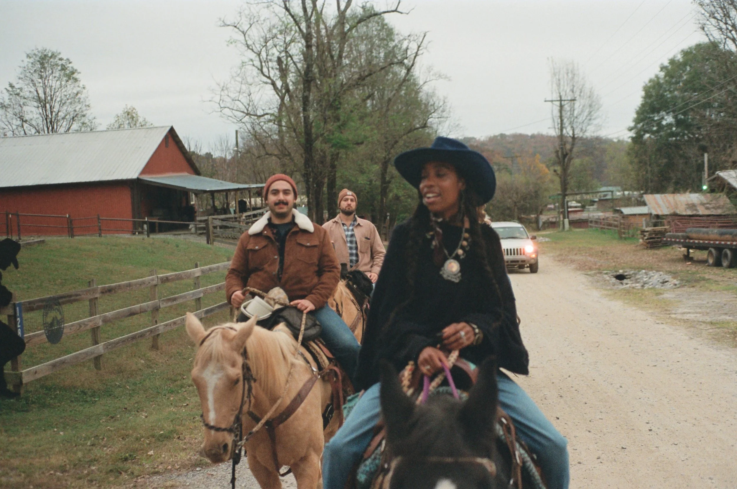 Three people riding horses along a rural gravel road with trees and farm buildings in the background, one woman wearing a large blue hat and black outfit, two men behind her.