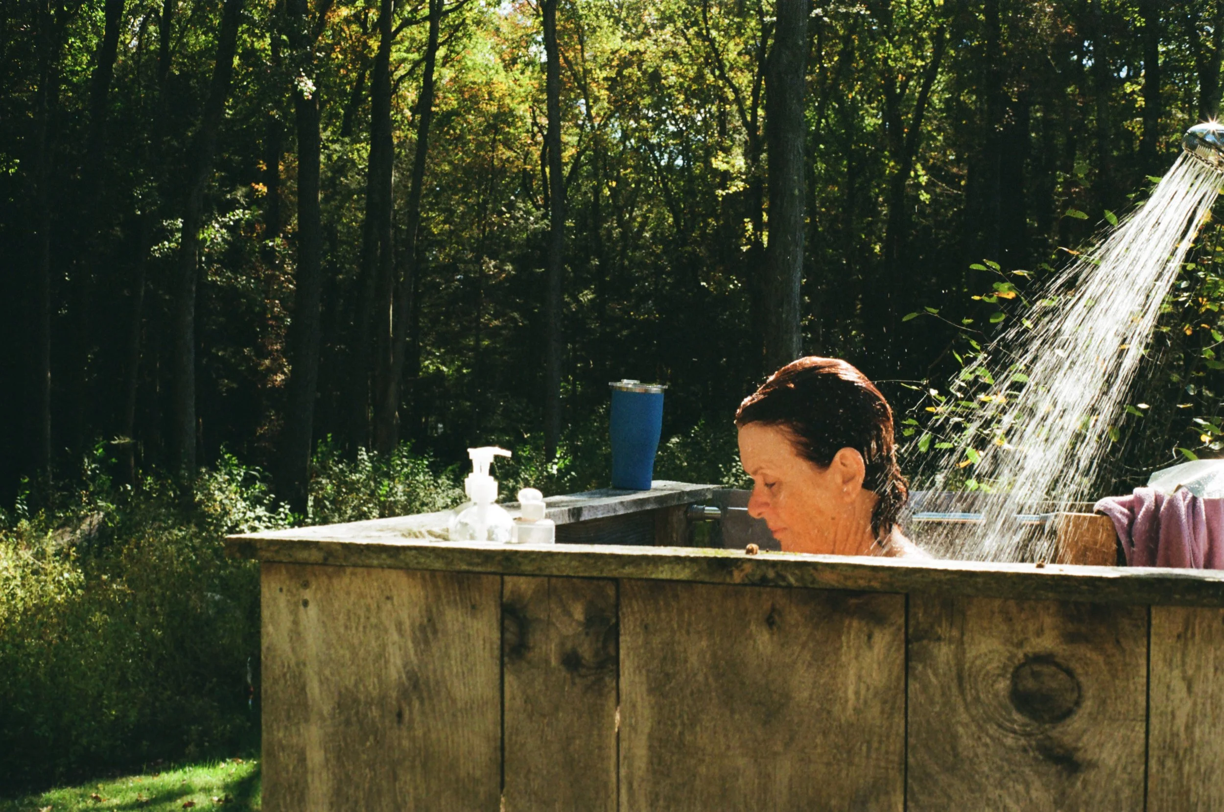 Woman taking a shower outdoors in a wooden tub surrounded by trees on a sunny day.