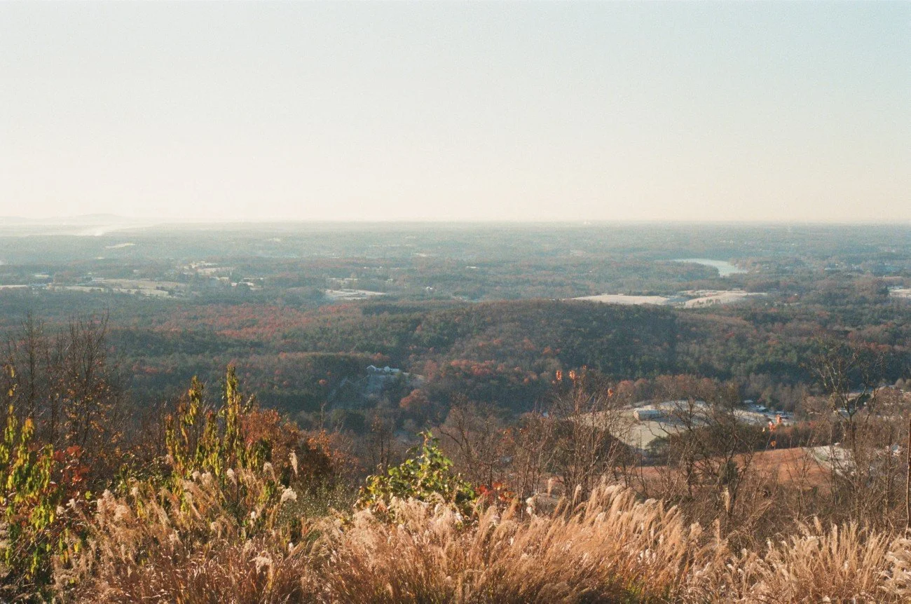 A wide landscape view from a hilltop showing rolling hills, trees with fall foliage, and a distant horizon under a clear sky.