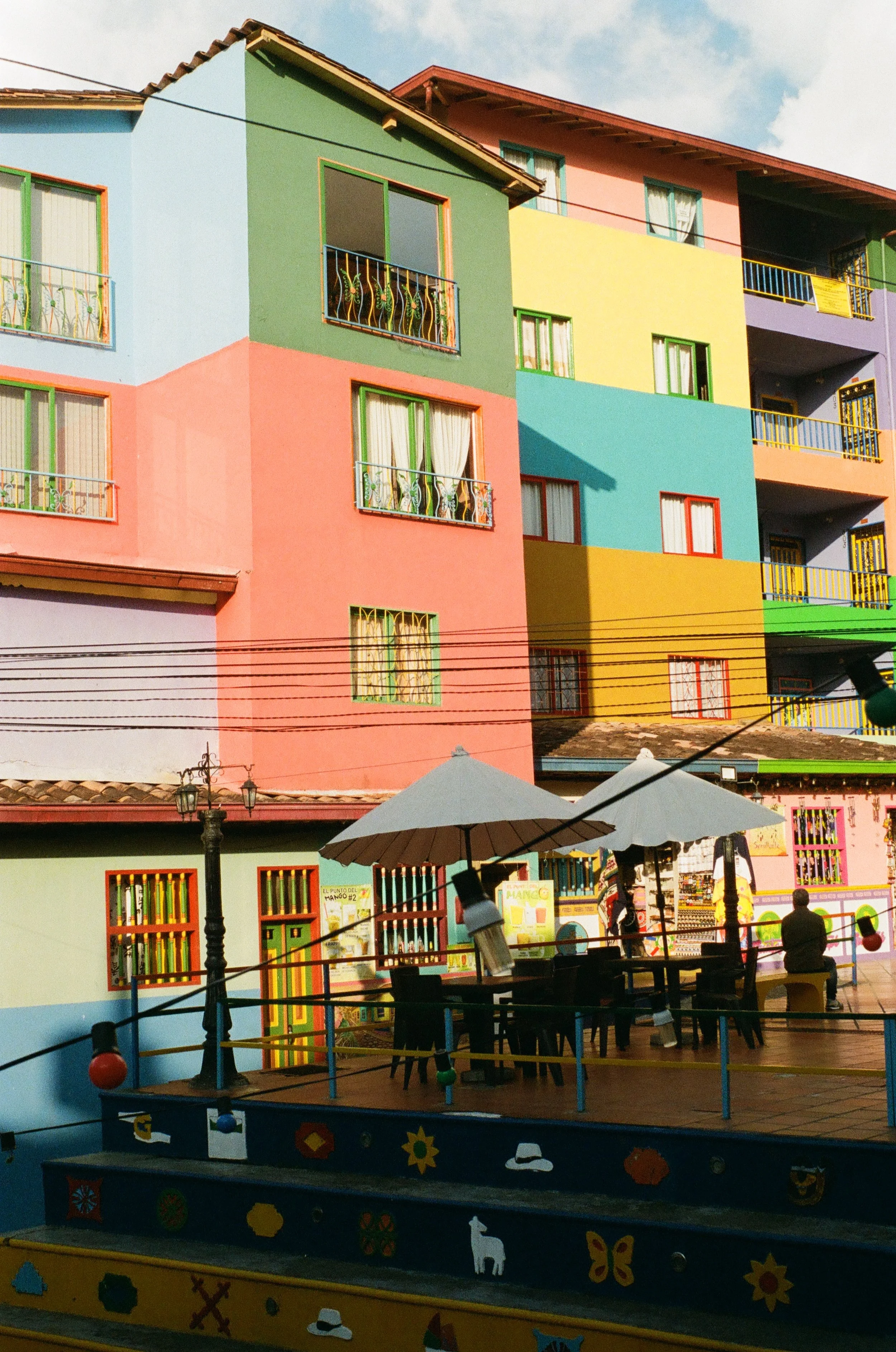 Colorful multi-story building with pink, green, blue, yellow, purple walls, and balconies with colorful railings; outdoor seating area with umbrellas and a person sitting, in a lively neighborhood.