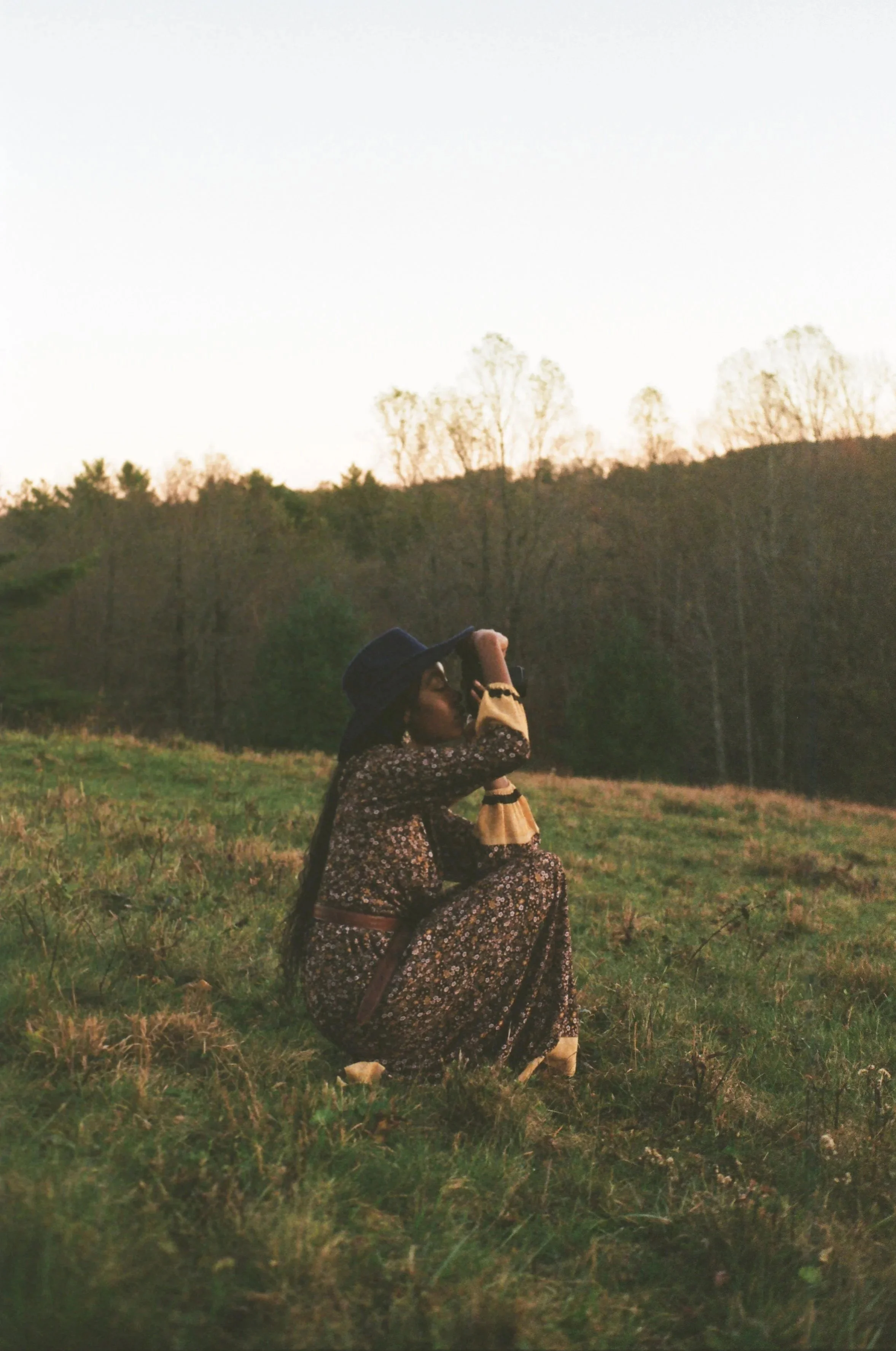 A woman kneeling in a field during sunset, wearing a floral dress, large hat, and taking a photograph with a camera.
