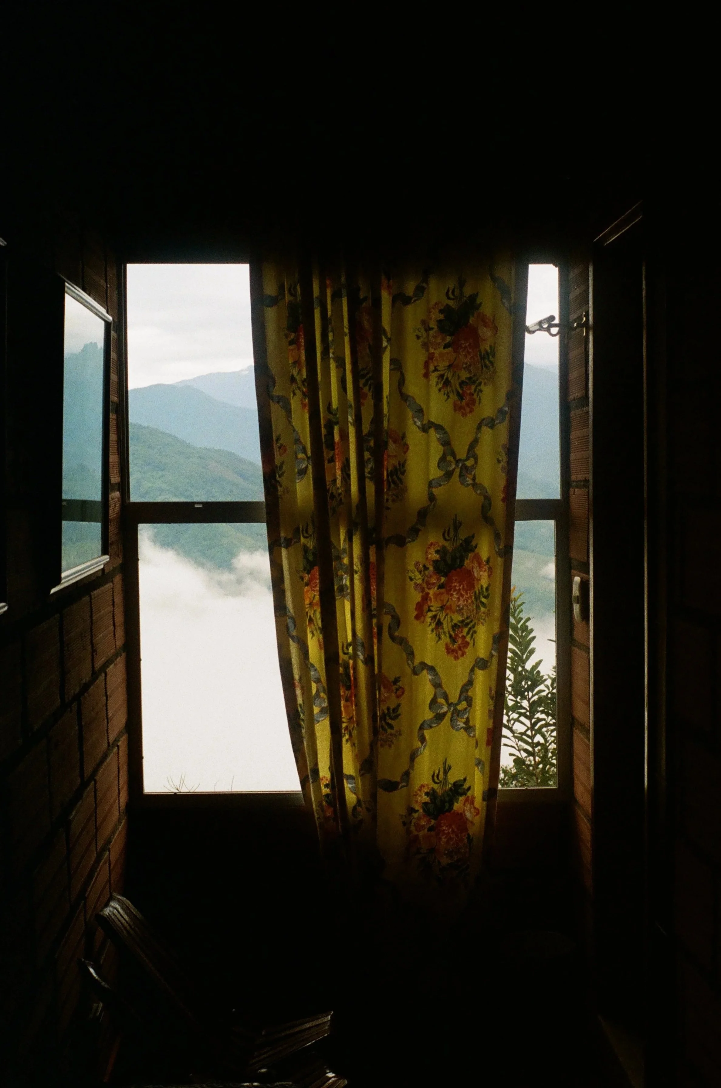 Open window with a yellow floral curtain revealing a misty mountain landscape.