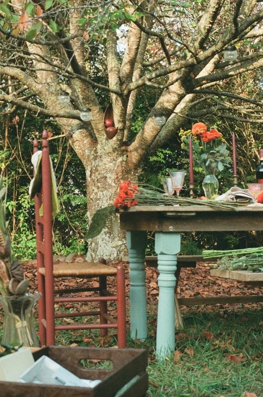 An outdoor table set under a large tree with a rustic wooden table, colorful flowers in vases, red candles, and vintage glassware, with autumn leaves on the ground and string lights hanging from the branches.