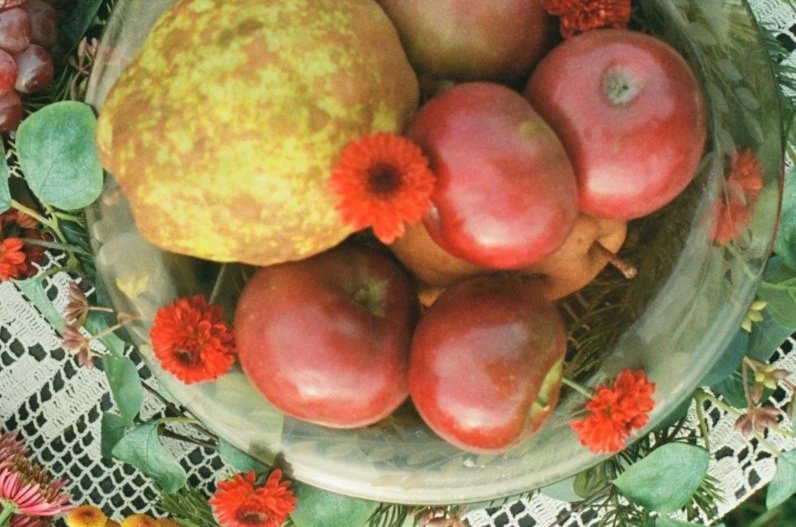 A glass bowl filled with five red apples, one yellow-green pear, and surrounded by small orange flowers and green leaves on a decorative lace tablecloth.