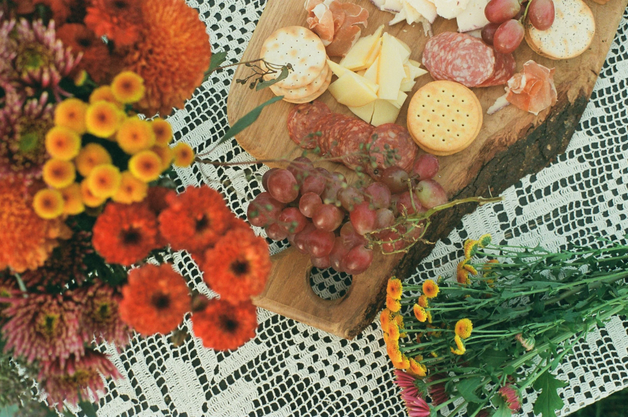 A cheese and charcuterie board with grapes, crackers, and slices of cheese on a rustic wooden serving tray, surrounded by colorful flowers on a lace tablecloth.